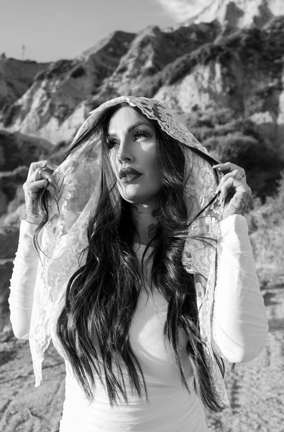 A woman with long dark hair and tattoos, wearing a lace shawl over her head, looking upward with a serious expression, standing outdoors with rocky mountains in the background.