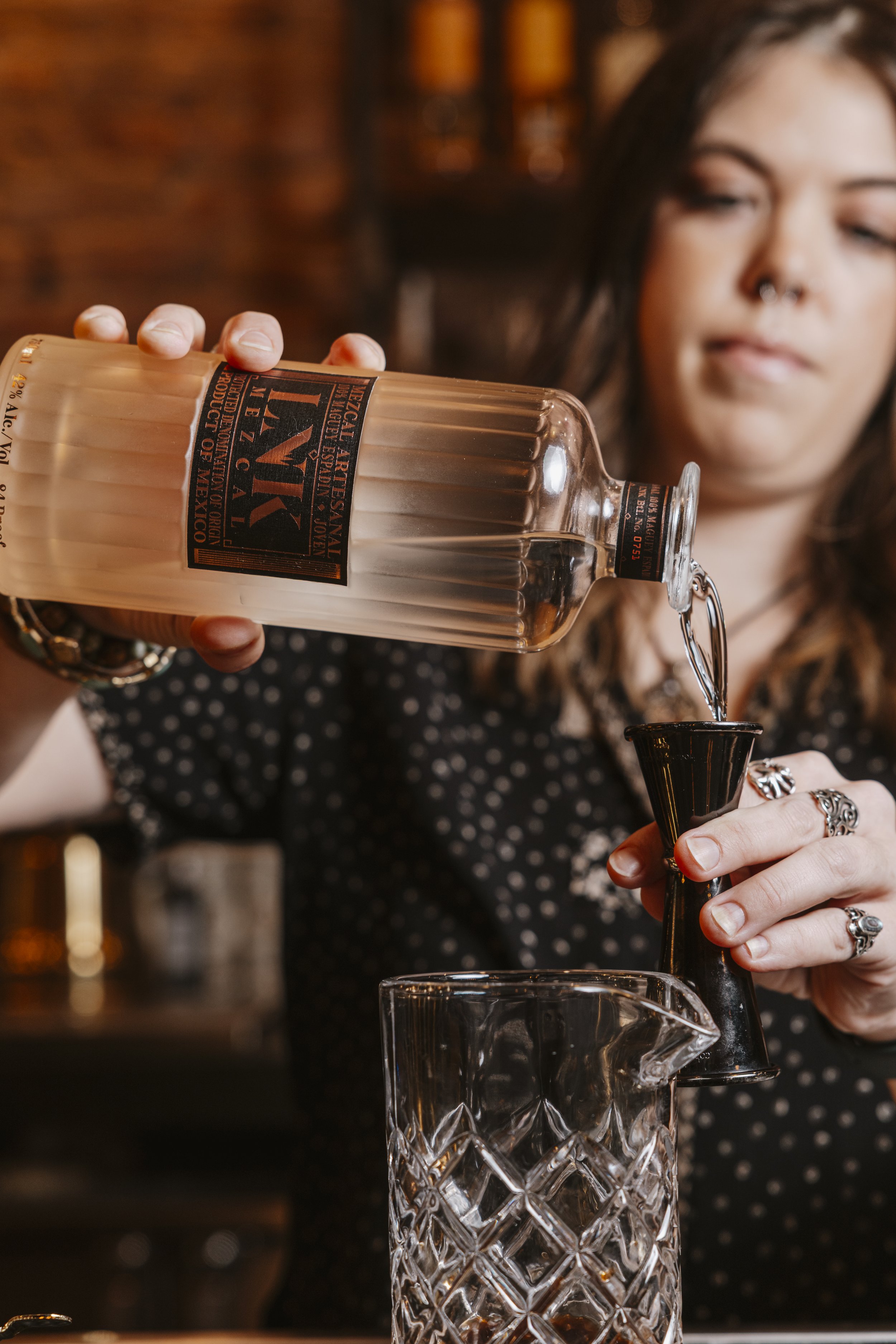 A woman with dark hair and jewelry pours liquor from a bottle into a jigger over a glass pitcher in a dimly lit bar or restaurant.