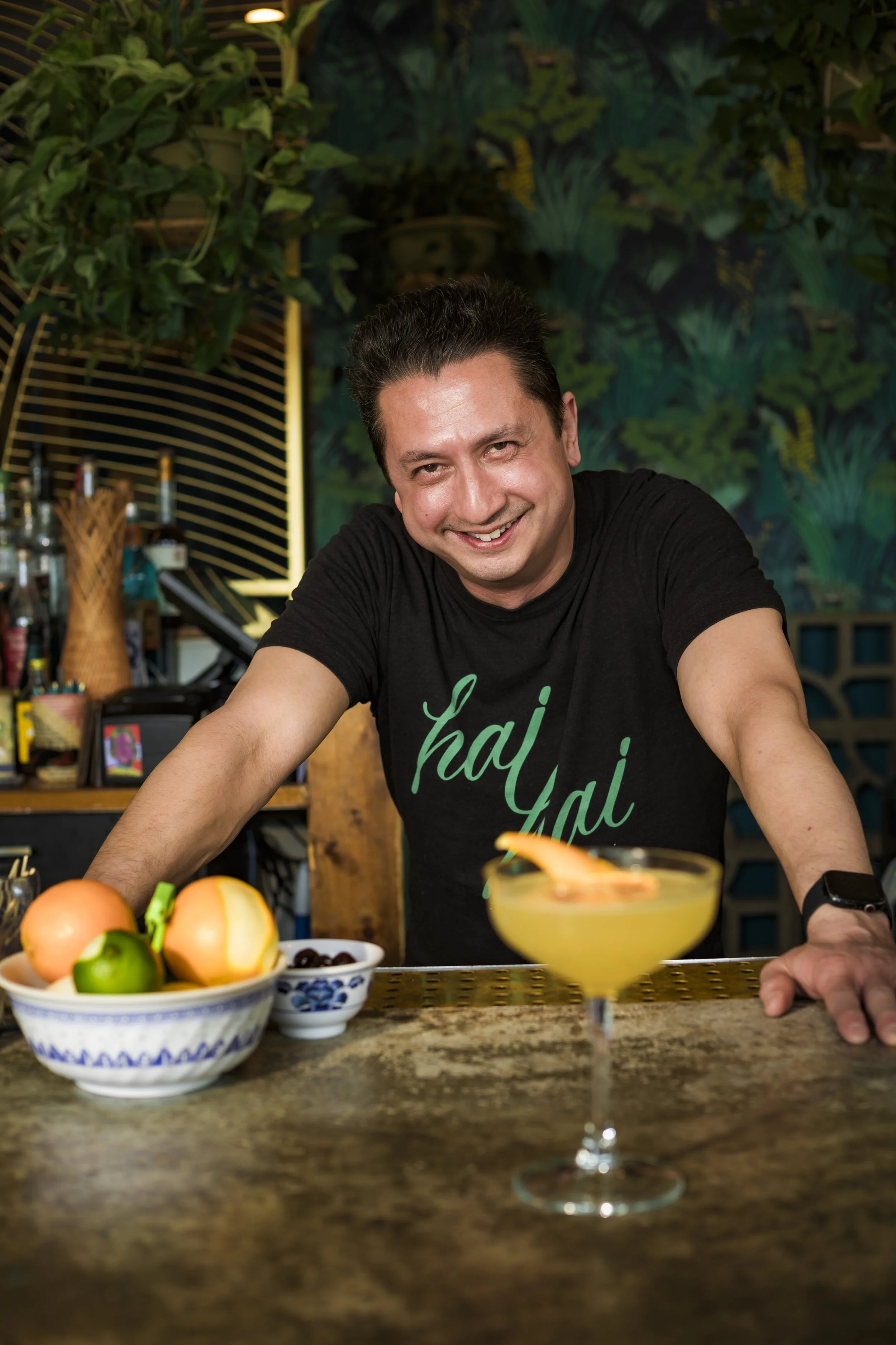 A man smiling at a bar counter with a cocktail glass in front of him, a bowl of fruit, and a small bowl of cherries. The background has green plants and decorative wall art.