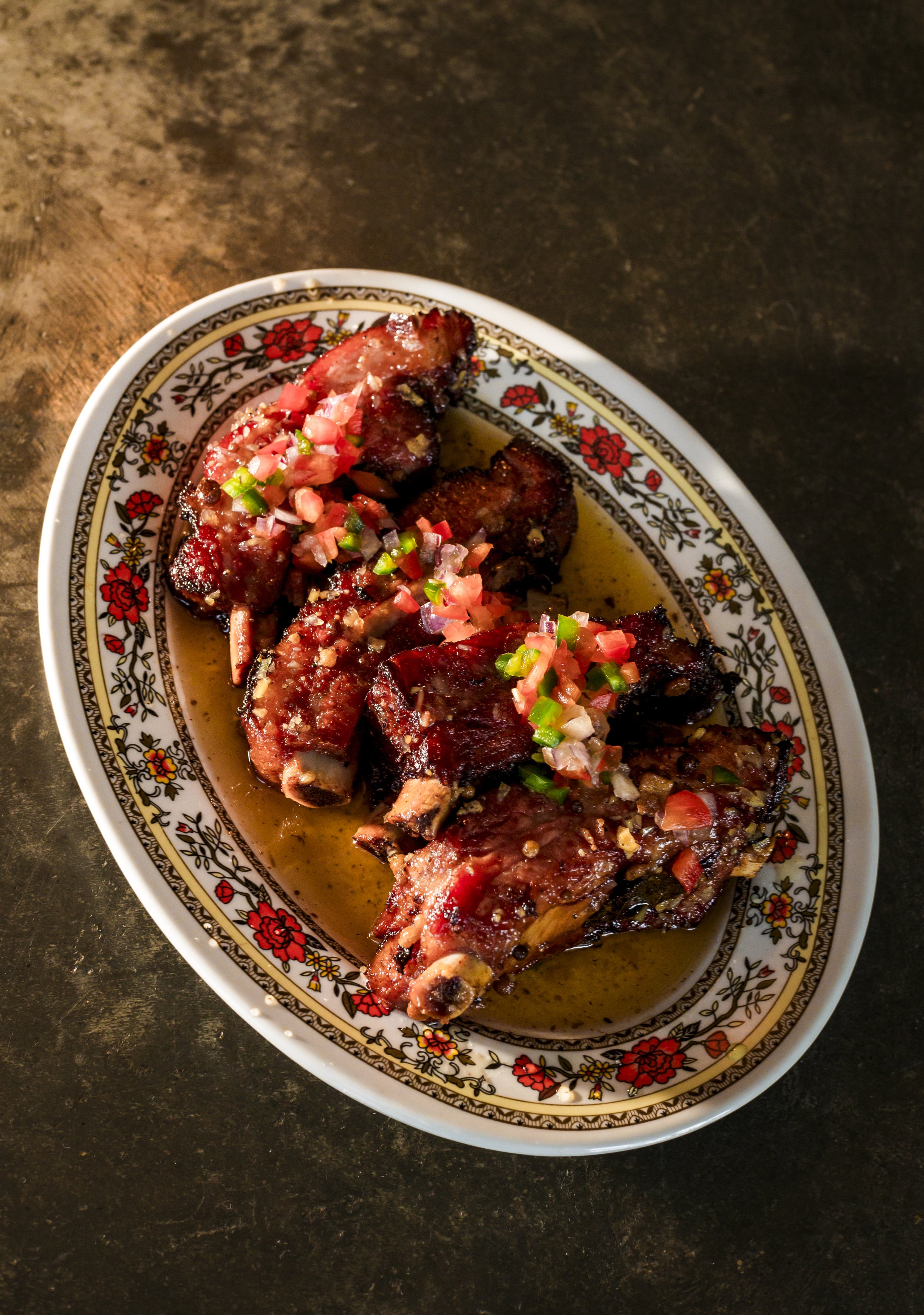 A decorative oval plate with grilled beef ribs topped with chopped tomatoes, onions, and green peppers, served in a flavorful sauce, on a dark wooden table.