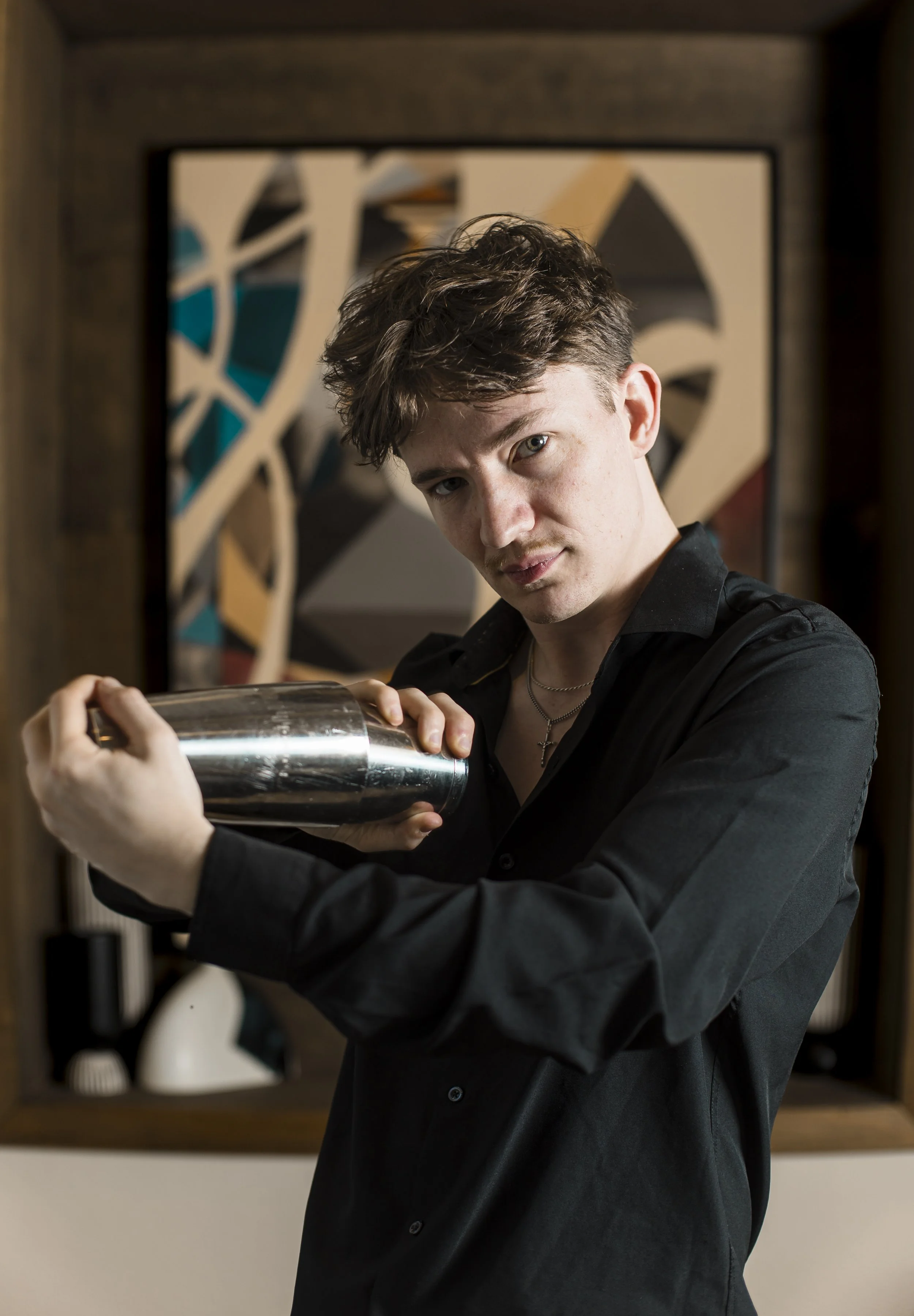 Young man with dark curly hair wearing a black shirt and silver cross necklace, holding a cocktail shaker, standing in front of abstract artwork.