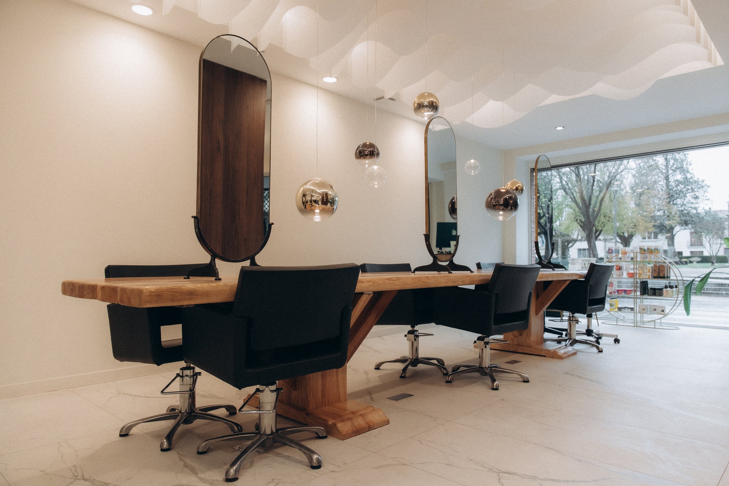 Salon de coiffure moderne avec trois stations de travail en bois massif, chaque équipée d'un grand miroir oval et d'une chaise noire, décor épuré avec suspension en formes organiques suspendues au plafond, grande fenêtre donnant sur une rue avec des 