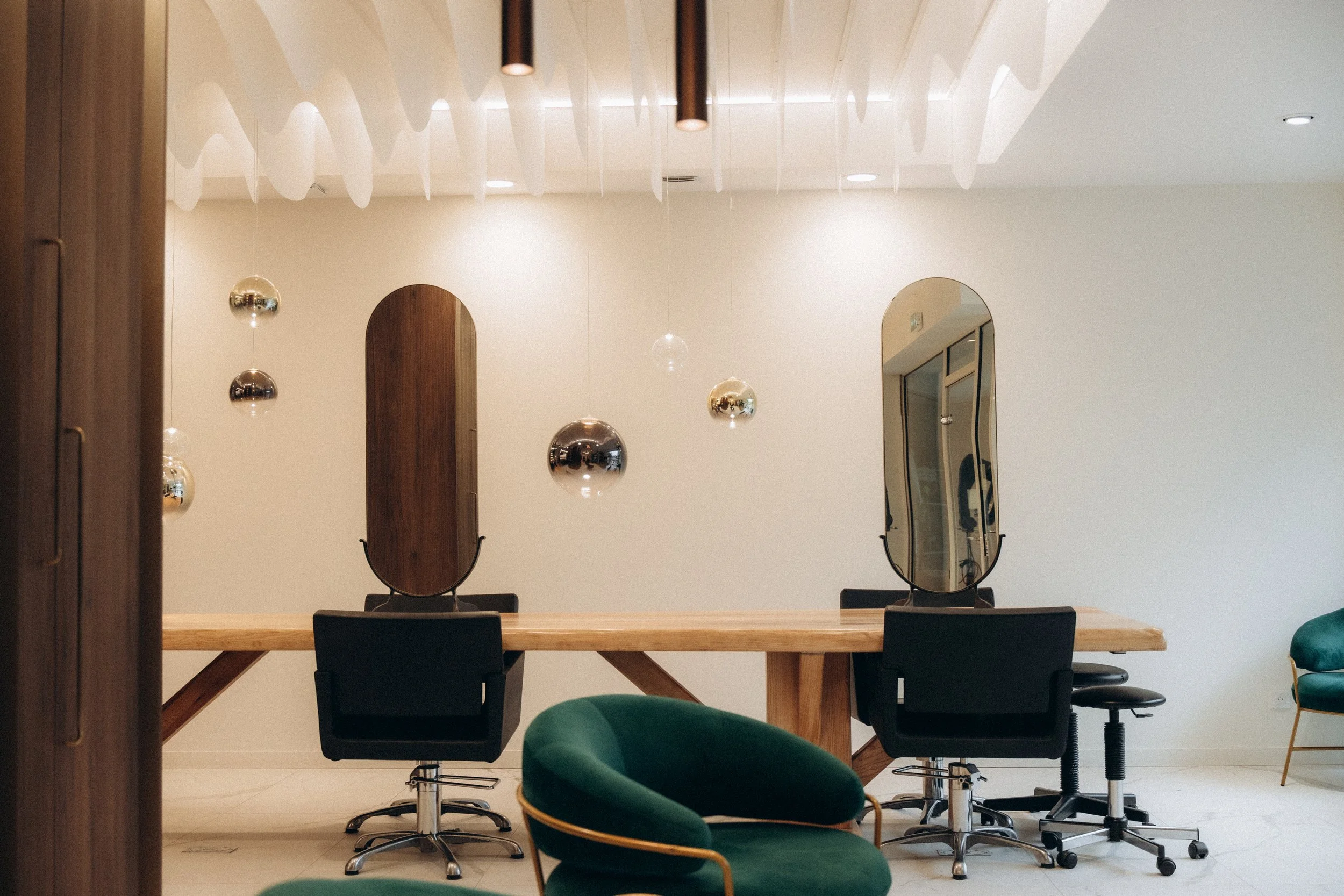 Salle de réunion moderne avec un grand tapis en bois, deux grands miroirs ovales, 6 chaises de bureau noires, 2 chaises vertes avec un cadre en métal doré, suspension de plusieurs ampoules suspendues en différents tons métalliques et un plafond blanc