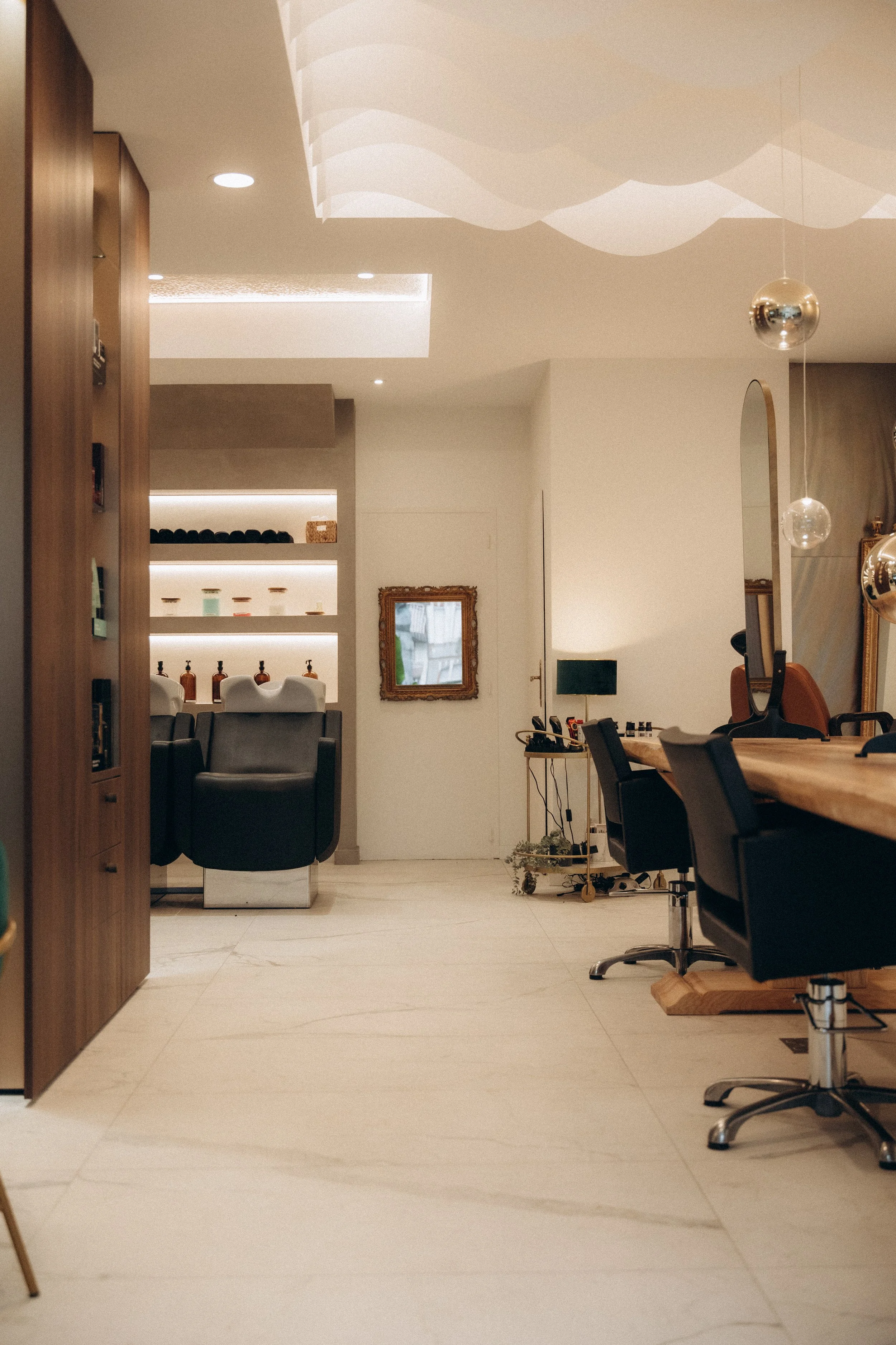 Intérieur d'un salon de coiffure ou spa avec des chaises de coiffure, un miroir, et un espace de détente avec des fauteuils et des étagères en arrière-plan.