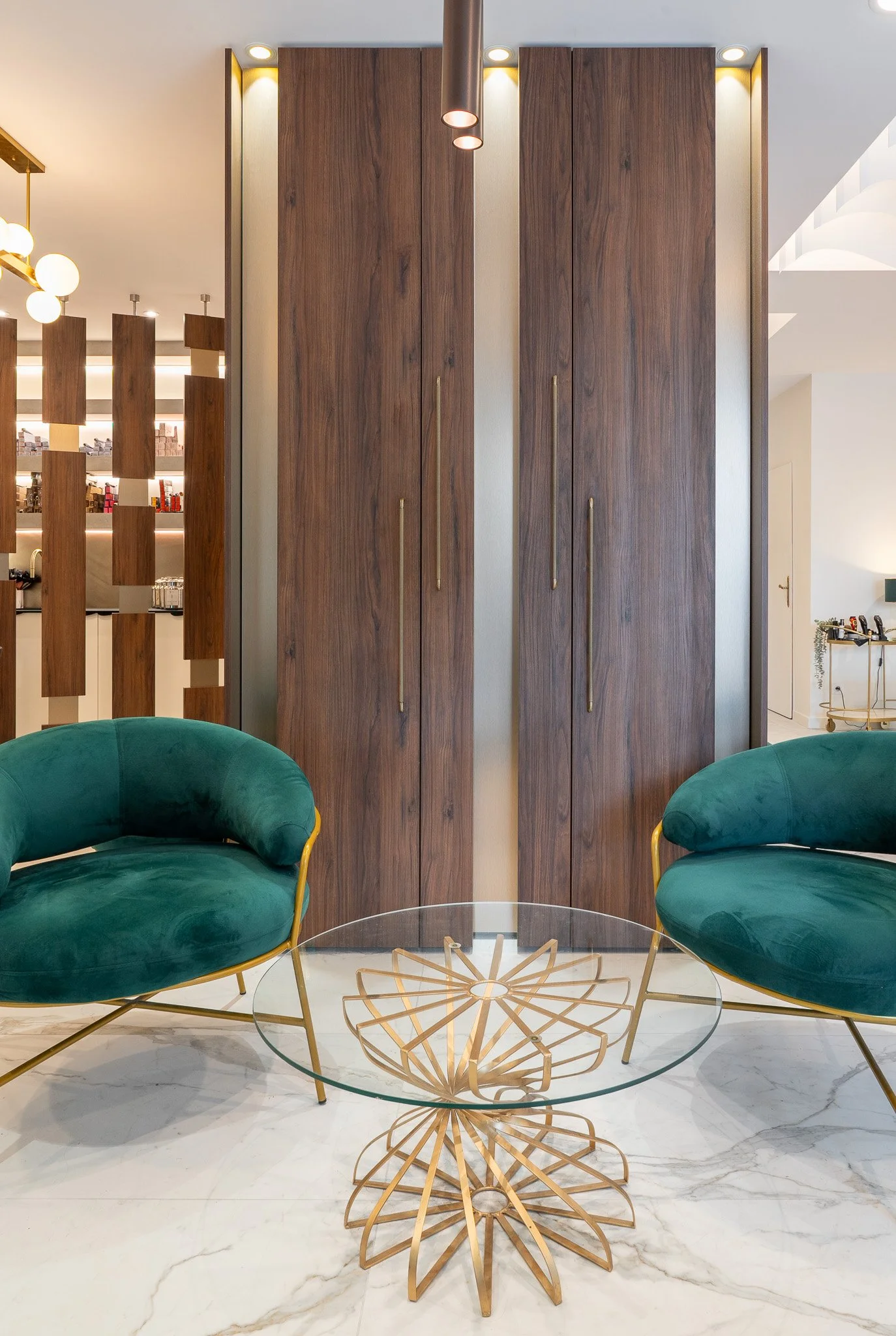 Une pièce avec deux fauteuils verts en velours, une table en verre avec un décor en bois en forme de papillon, un grand placard en bois derrière, et un sol en marbre blanc.