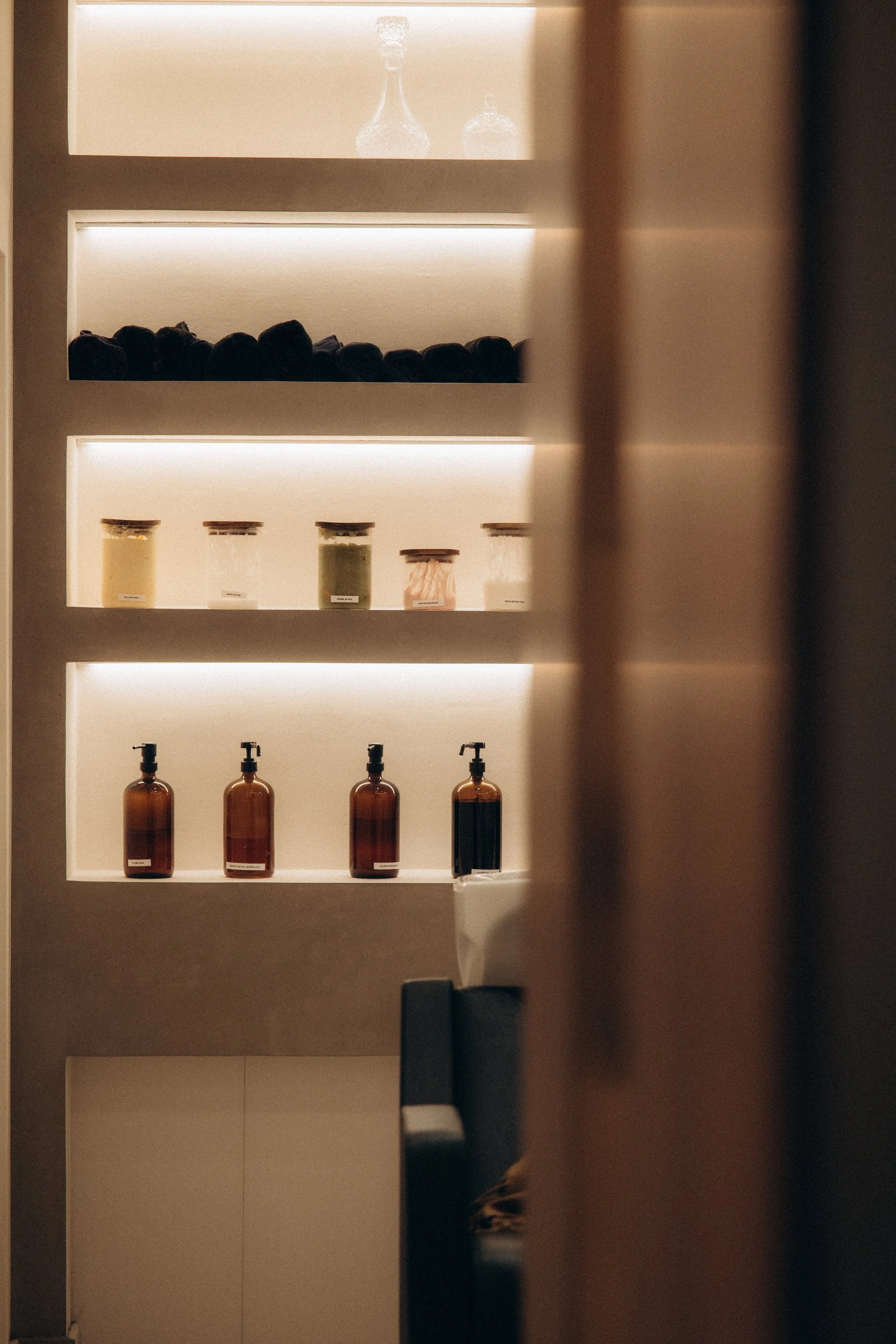 Étagère éclairée contenant des bocaux, bouteilles en plastique avec pompe, et autres contenants derrière une porte entrouverte.