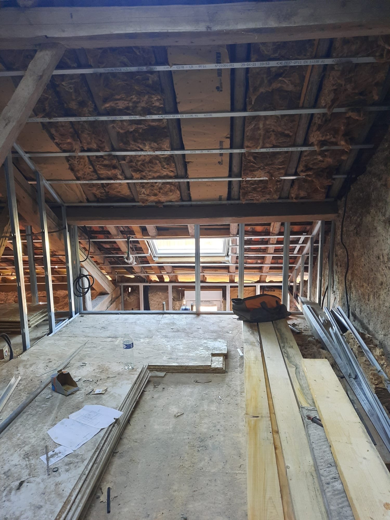 Intérieur d'un espace en construction avec ossature en métal, poutres en bois, isolation en laine de roche, fenêtre de toit, et des matériaux de construction éparpillés.