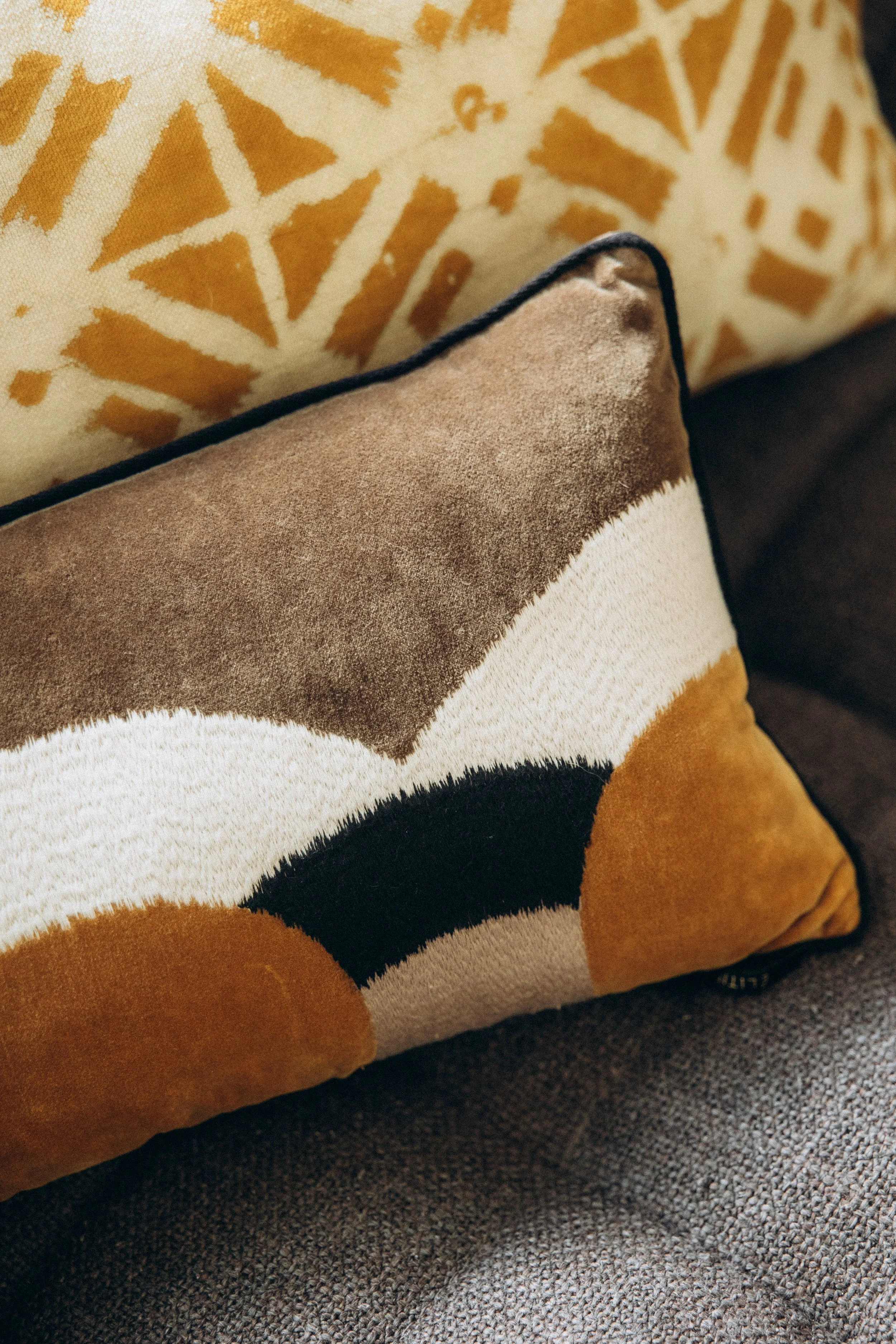 Coussins décoratifs sur un canapé, un en tissu beige, un autre en velours marron, et un avec un motif géométrique noir, blanc, beige et marron.