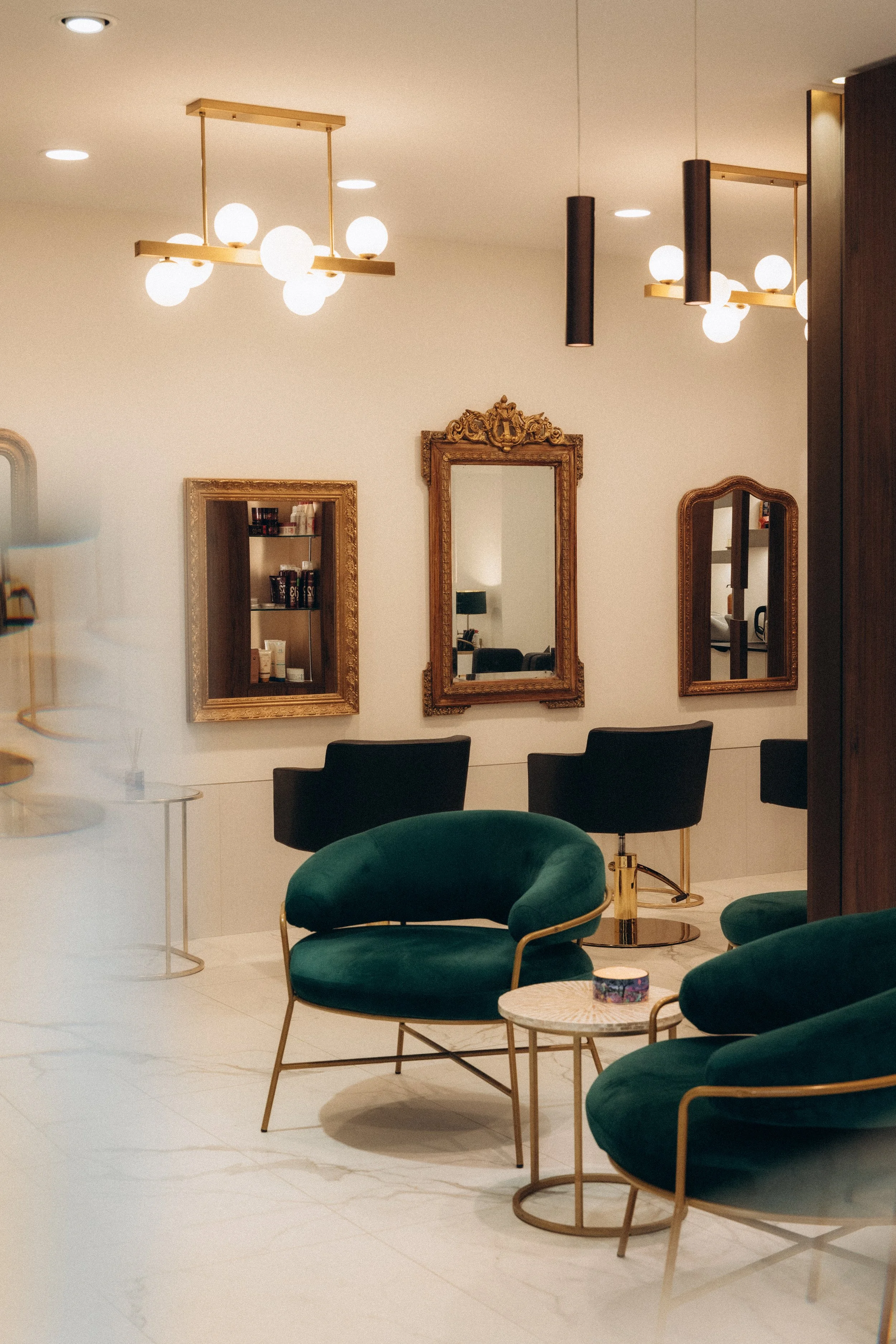 Un salon avec des fauteuils en velours vert, des miroirs anciens en cadres dorés, des lampes suspendues modernes, et une décoration élégante.