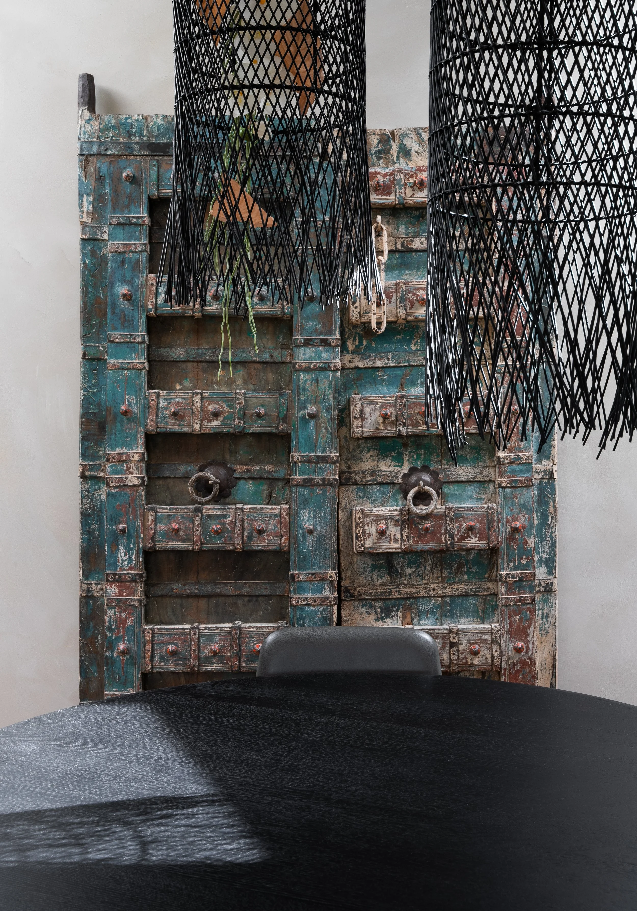 Deux grands luminaires noirs en forme de lamelles suspendus au plafond, au-dessus d'une table noire. En arrière-plan, un meuble ancien en bois avec une peinture bleu délavée, orné de quincaillerie en fer rouillé.