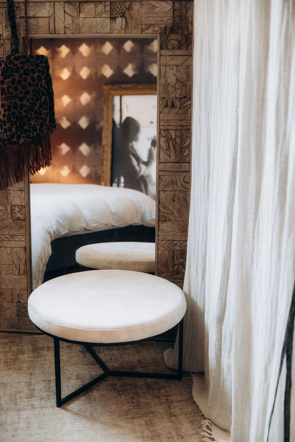 Un miroir dans une chambre reflète une silhouette de femme assise, avec un lit en arrière-plan, des rideaux clairs et un mur en bois orné de motifs.