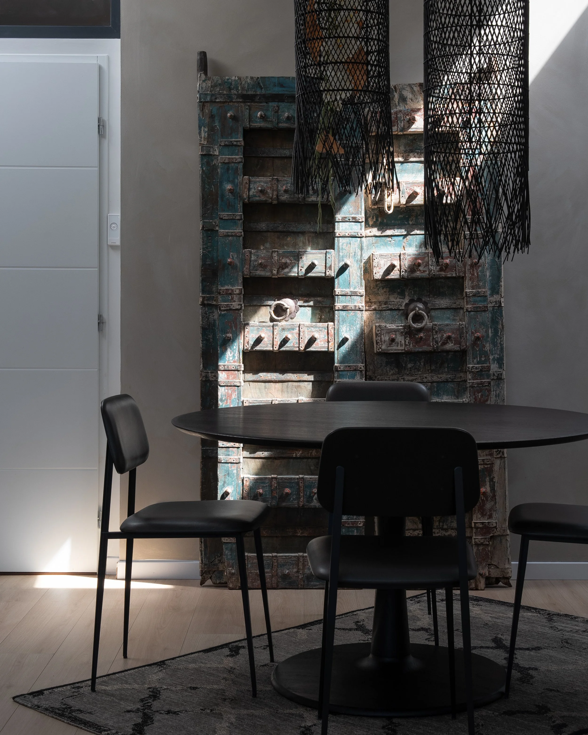 Une table ronde noire entourée de trois chaises en fauteuil en cuir noir dans une pièce moderne. En arrière-plan, un mur décoratif en bois vieux et une suspension noire en forme de parasol. La lumière naturelle éclaire partiellement la pièce.