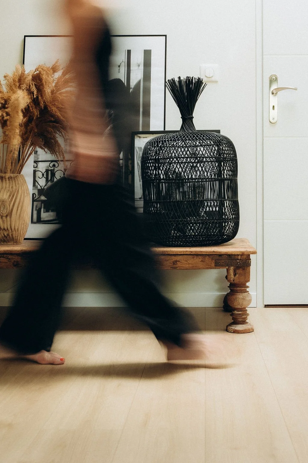 Un intérieur avec un meuble en bois, une grande jarre en bois avec des plantes, une affiche encadrée, et un vase noir en rotin. Personne passant devant, floue, en marchant.