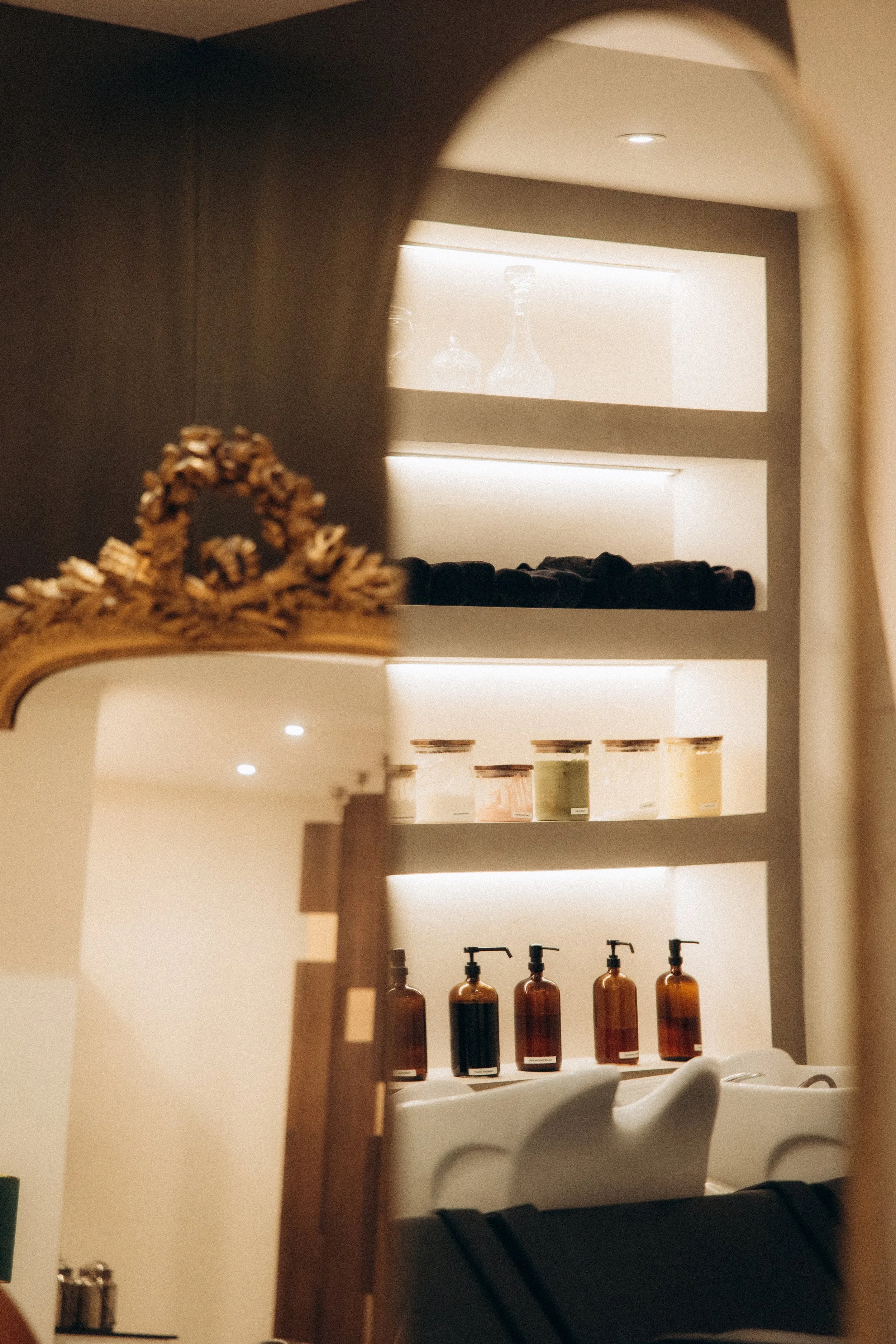 Un miroir reflète une étagère avec des bouteilles de savon ou de lotion, des bocaux, et un espace de lavage de cheveux ou de spa dans une salle de bain ou un salon de beauté.