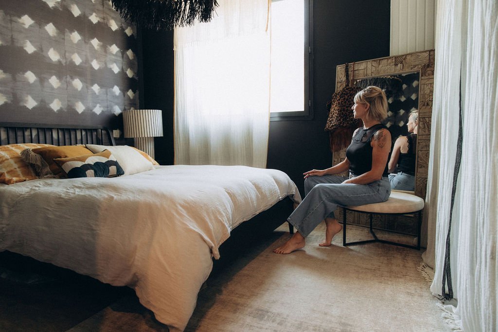 Une femme assise sur une chaise dans une chambre, regardant vers la fenêtre. La chambre a un lit avec des coussins, une lampe de chevet, un miroir en bois et des rideaux blancs.