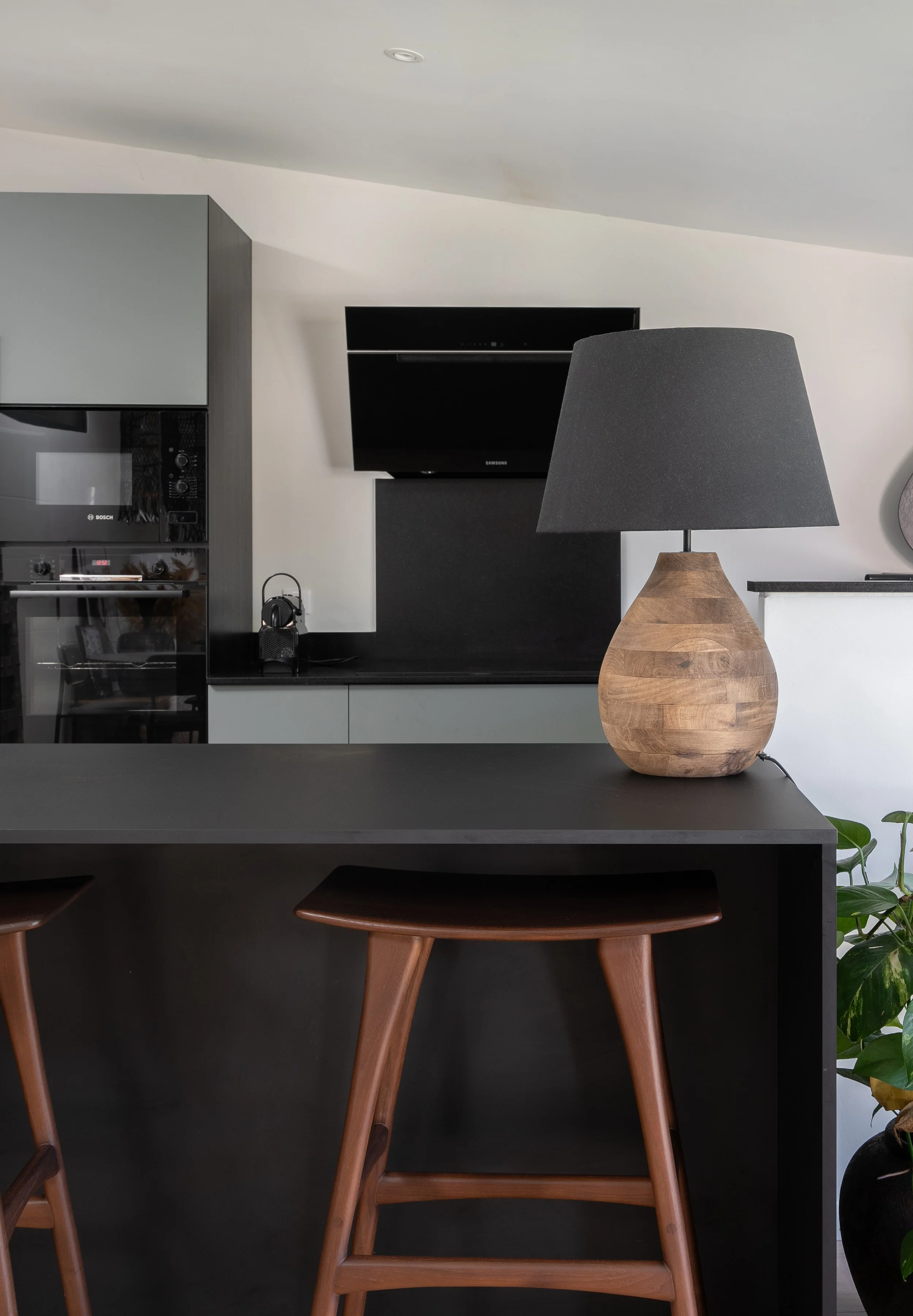Intérieur de cuisine moderne avec un éclairage doux, table noire, lampe en bois à abat-jour gris, plantes vertes en pot, et appareils électroménagers encastrés.