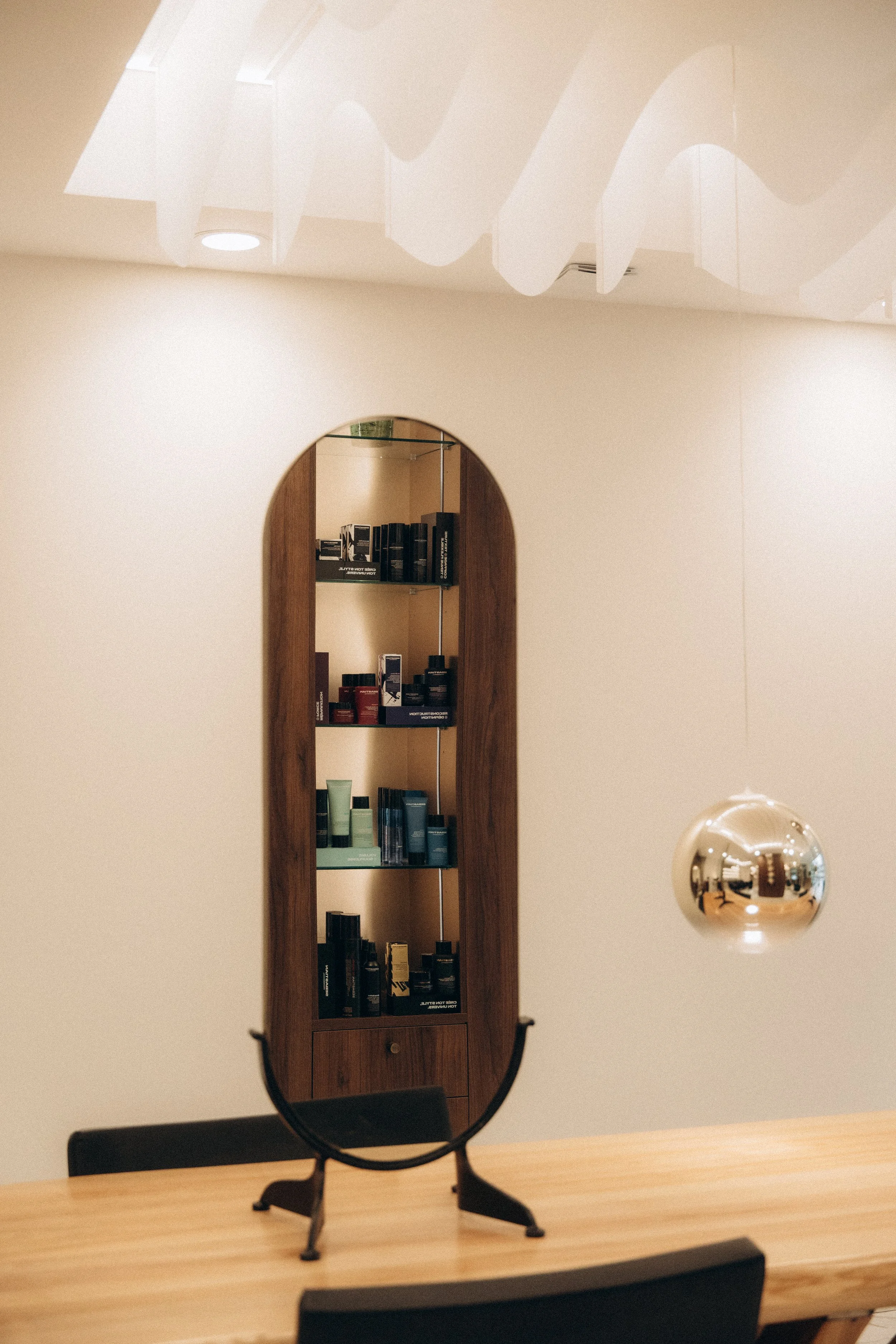 Miroir de table avec meuble en bois derrière, réflexions sur le meuble et la pièce, lampe suspendue métallique ronde, plafond blanc avec lumières encastrées.