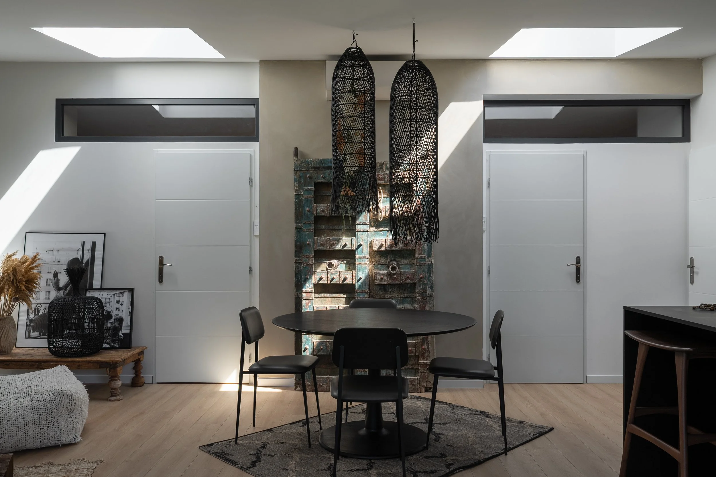 Salle à manger moderne avec une table ronde noire, quatre chaises noires, deux suspensions en osier noir, un mur en briques peintes, deux portes blanches, et un bureau en bois comme décoration.