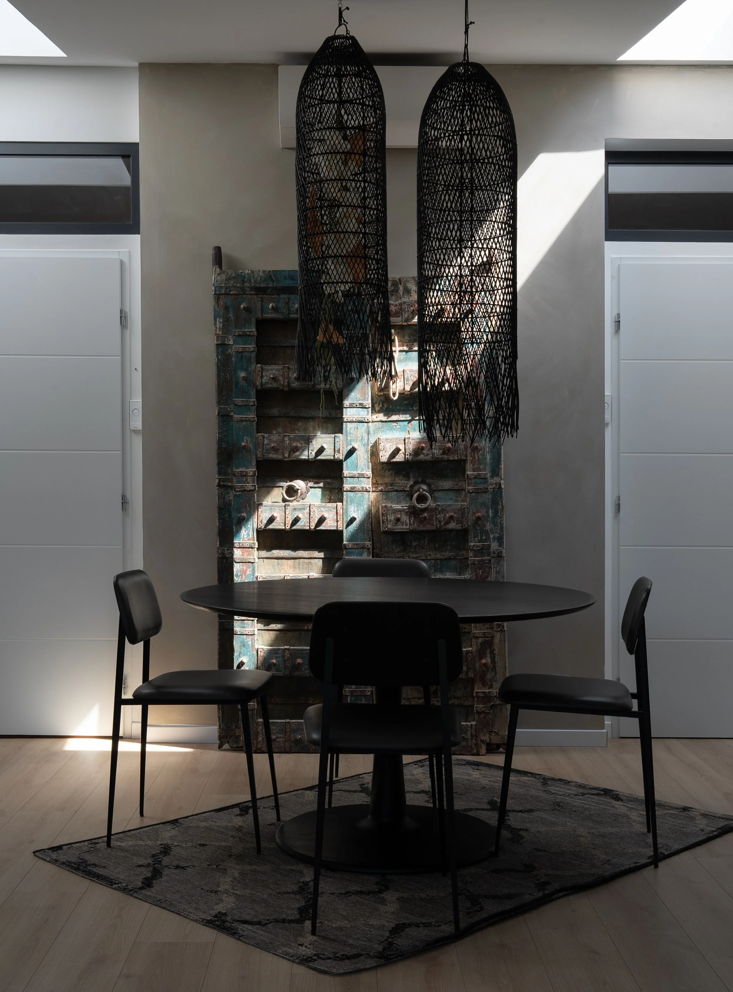 Une salle à manger avec une table ronde noire entourée de quatre chaises noires, un tapis sous la table, deux lampes suspendues en rotin noir, et une pièce de décoration murale en bois et métal dans le fond.