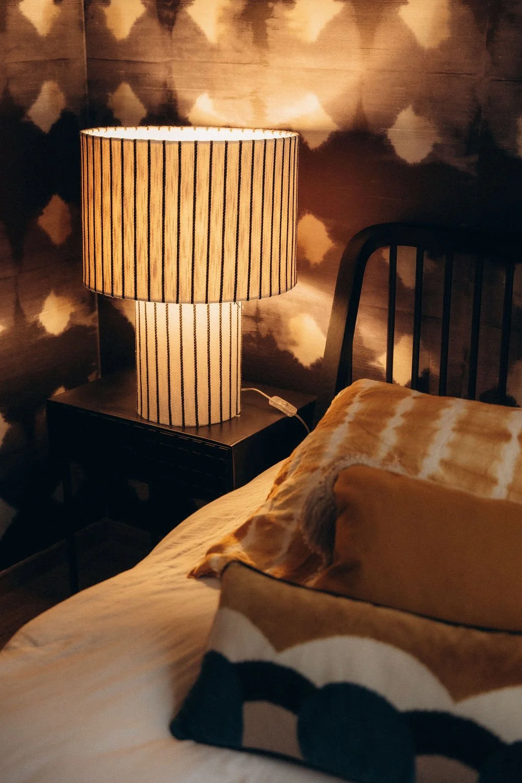 Lampe de chevet à rayures noires et beiges allumée à côté d'un lit avec oreillers dans une chambre au mur avec motif nuages.