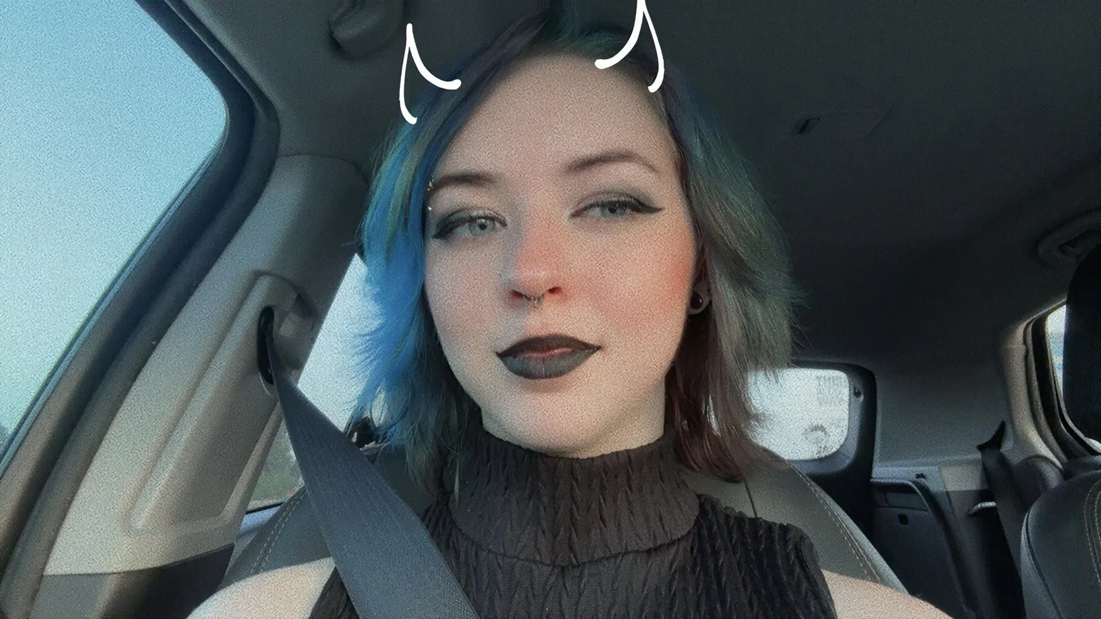 A woman with blue and green hair, wearing black lipstick and a septum ring, sitting in a car and taking a selfie.