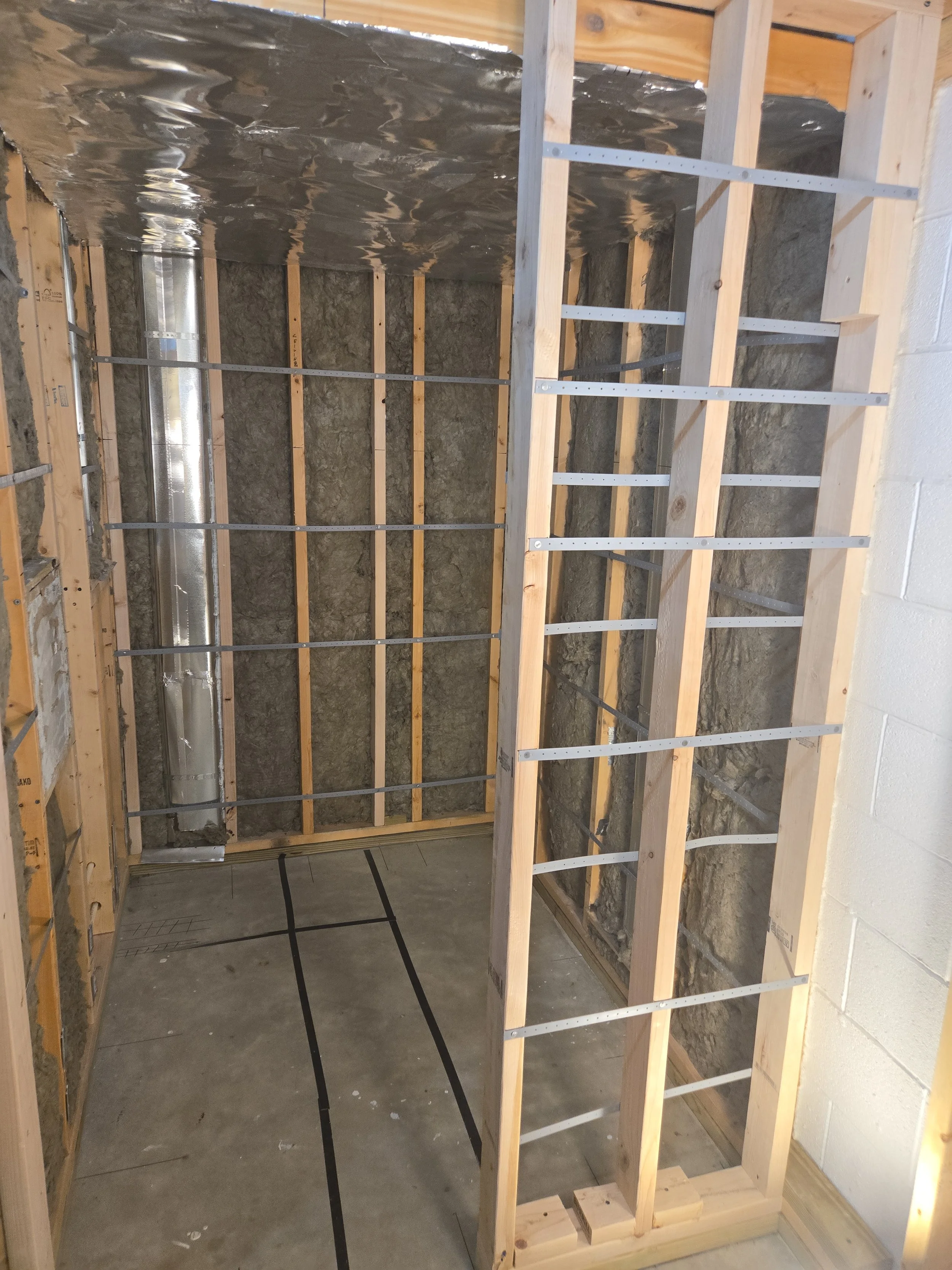 Partially constructed room with wooden framing and insulation, concrete floor, and metallic ceiling lining.