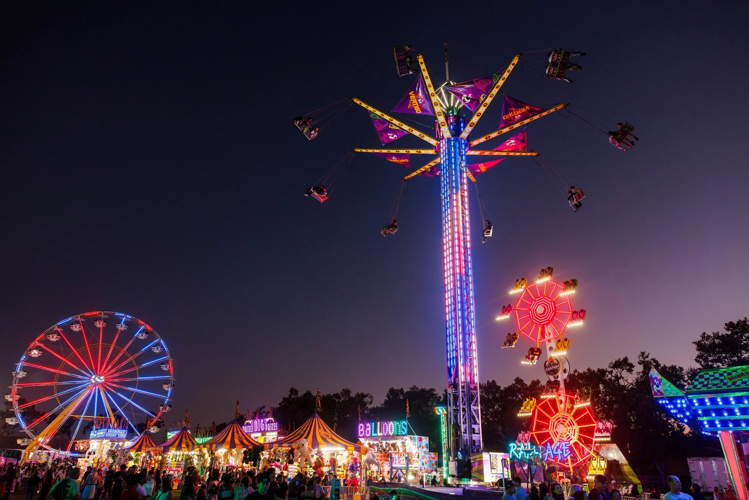 Brightly lit amusement park at night featuring a large swing ride in the air, a colorful Ferris wheel on the left, and a smaller carousel and game booths in the foreground, with people enjoying the fair.