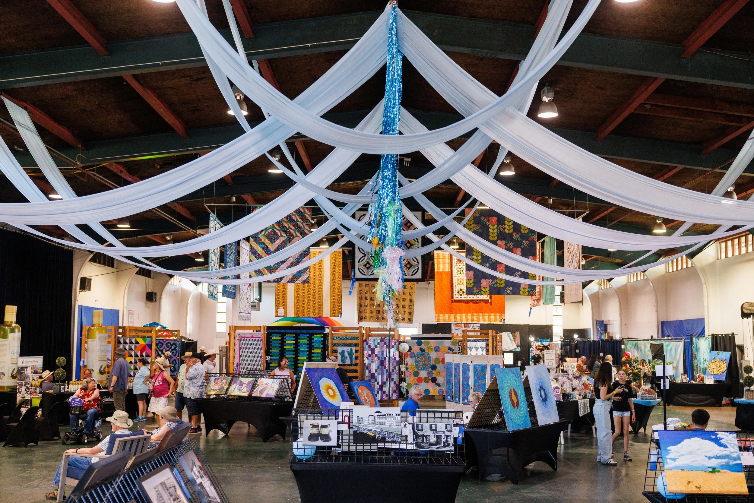 Indoor art fair with various booths displaying colorful paintings and crafts. White fabric drapes decorate the ceiling, and quilts are hanging on the walls. People are browsing and interacting with vendors.