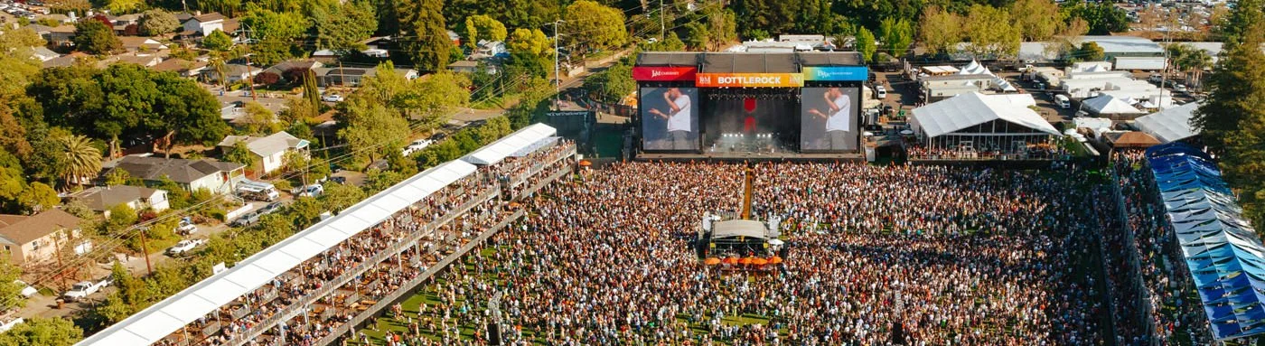 A large outdoor music concert with a massive crowd gathered in front of a stage with two large screens, surrounded by tents and trees.