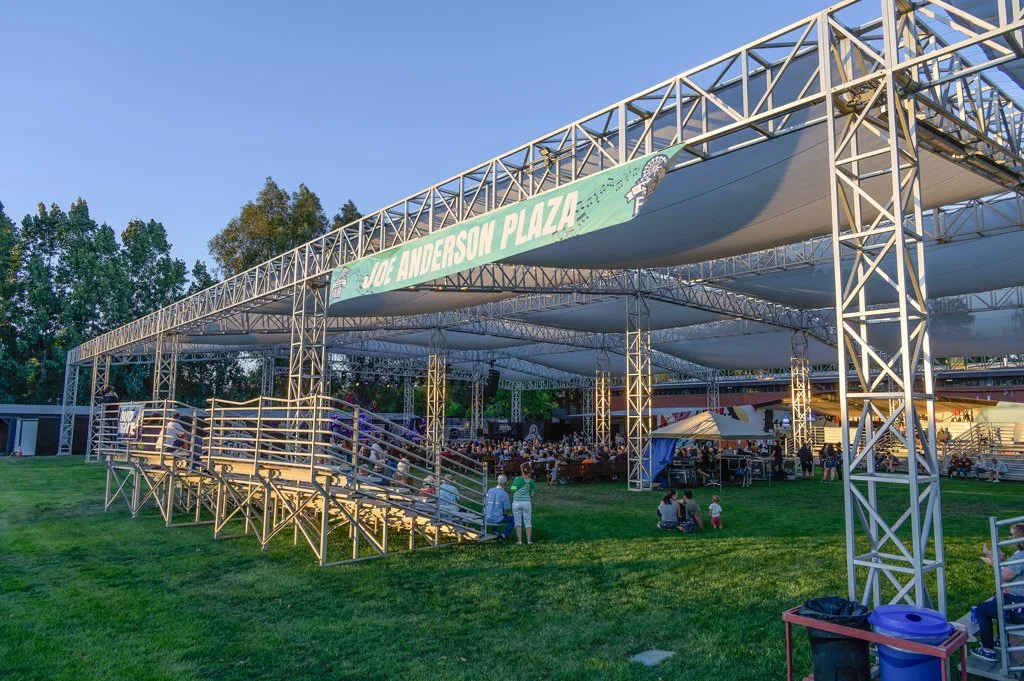 Outdoor concert at Joe Anderson Plaza with a stage, people sitting on grass, and trees in the background during early evening.