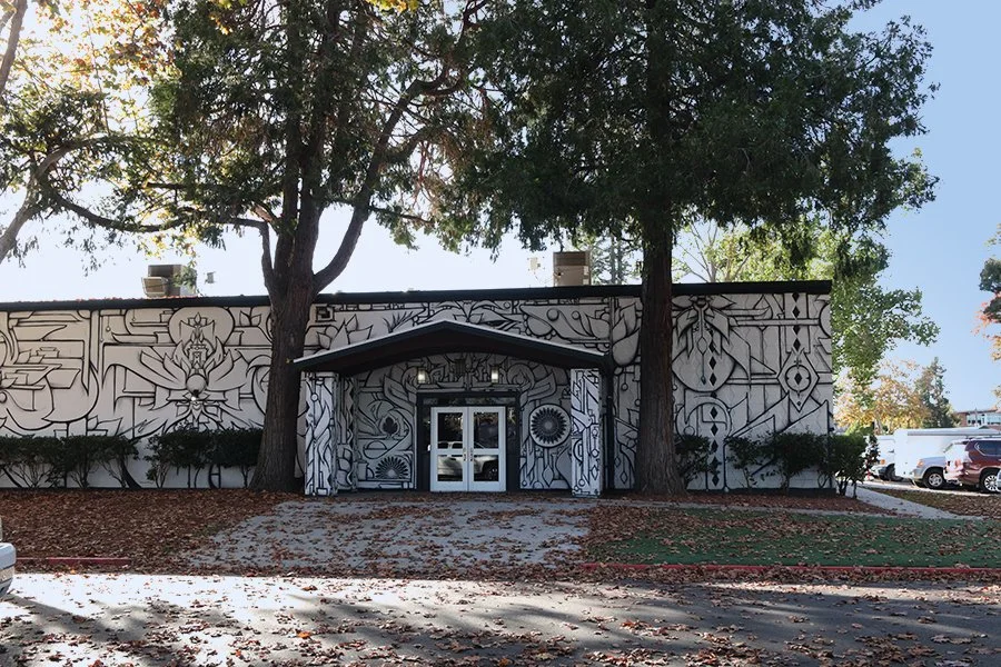 A building with a mural on the exterior wall, featuring black and white abstract and geometric designs, situated under large trees on a sunny day.