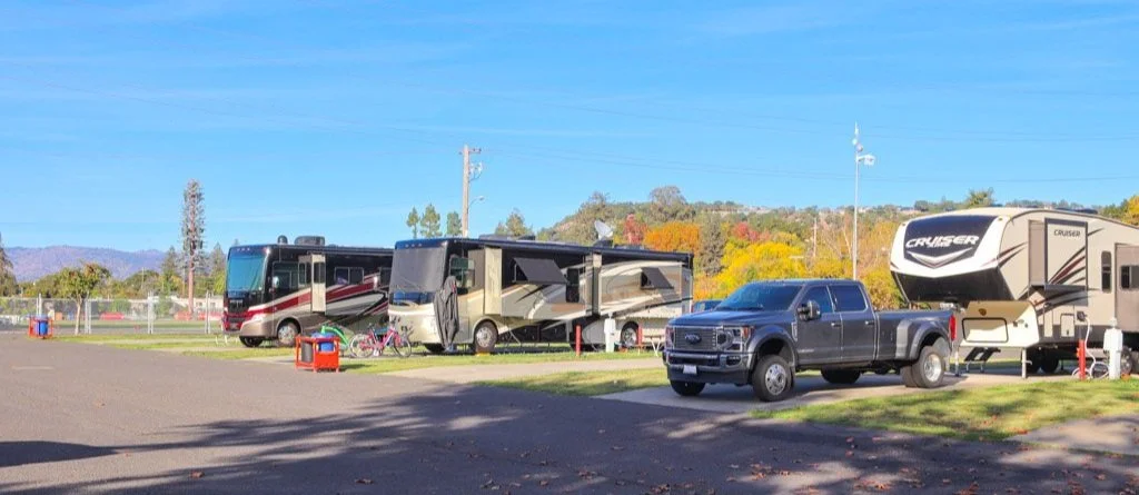Several recreational vehicles (RVs) and a pickup truck parked at a campsite or RV park under a clear blue sky, with trees and hills in the background.