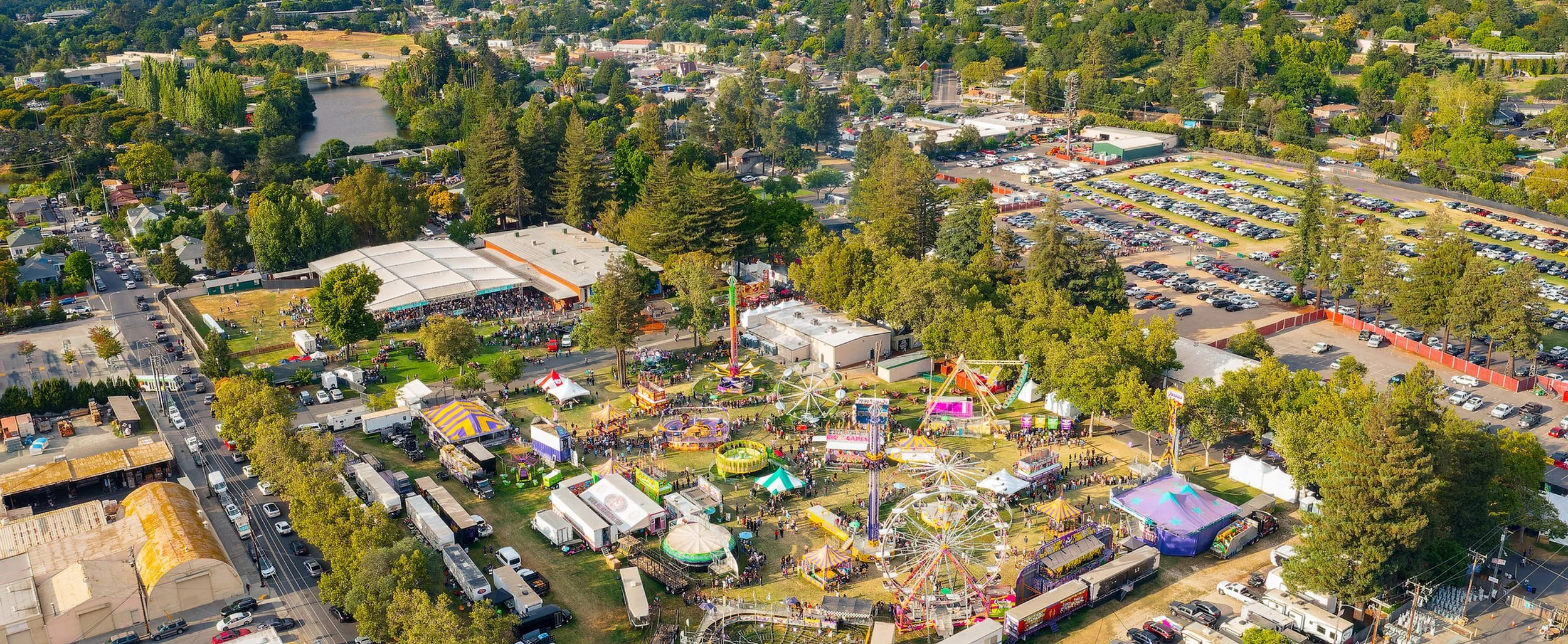 An aerial view of a fairground with rides, food stalls, and many visitors, surrounded by trees, buildings, and a parking lot.