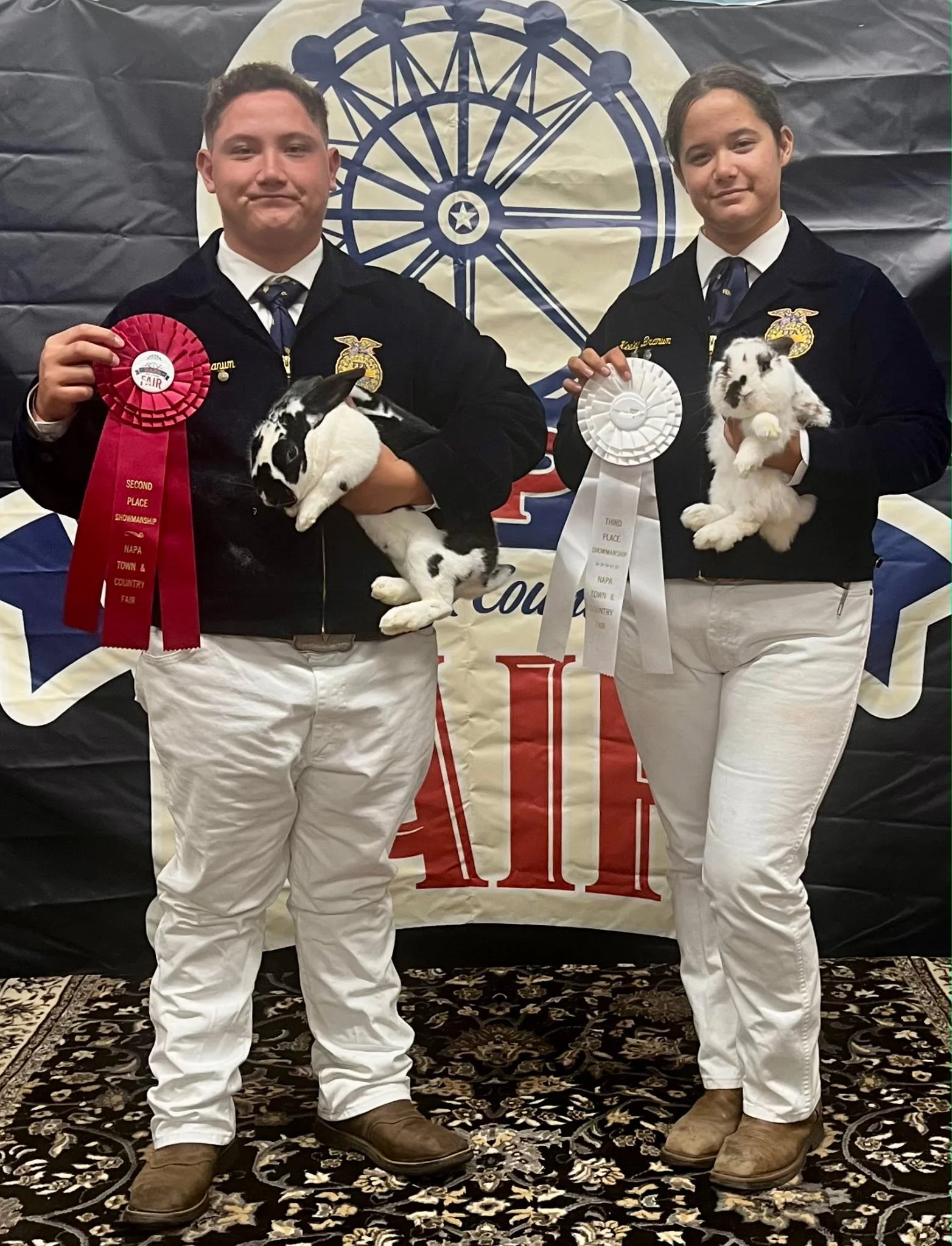 Two young people in FFA jackets standing on a decorative rug in front of a backdrop with a Ferris wheel design. Each is holding a rabbit and a ribbon indicating awards from a fair or show, with the person on the left holding a black and white rabbit 