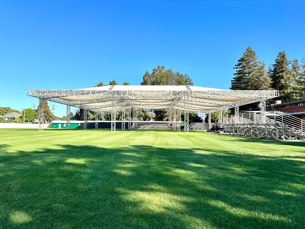 Large outdoor stage with white canopy on a grassy field, surrounded by trees and a clear blue sky.