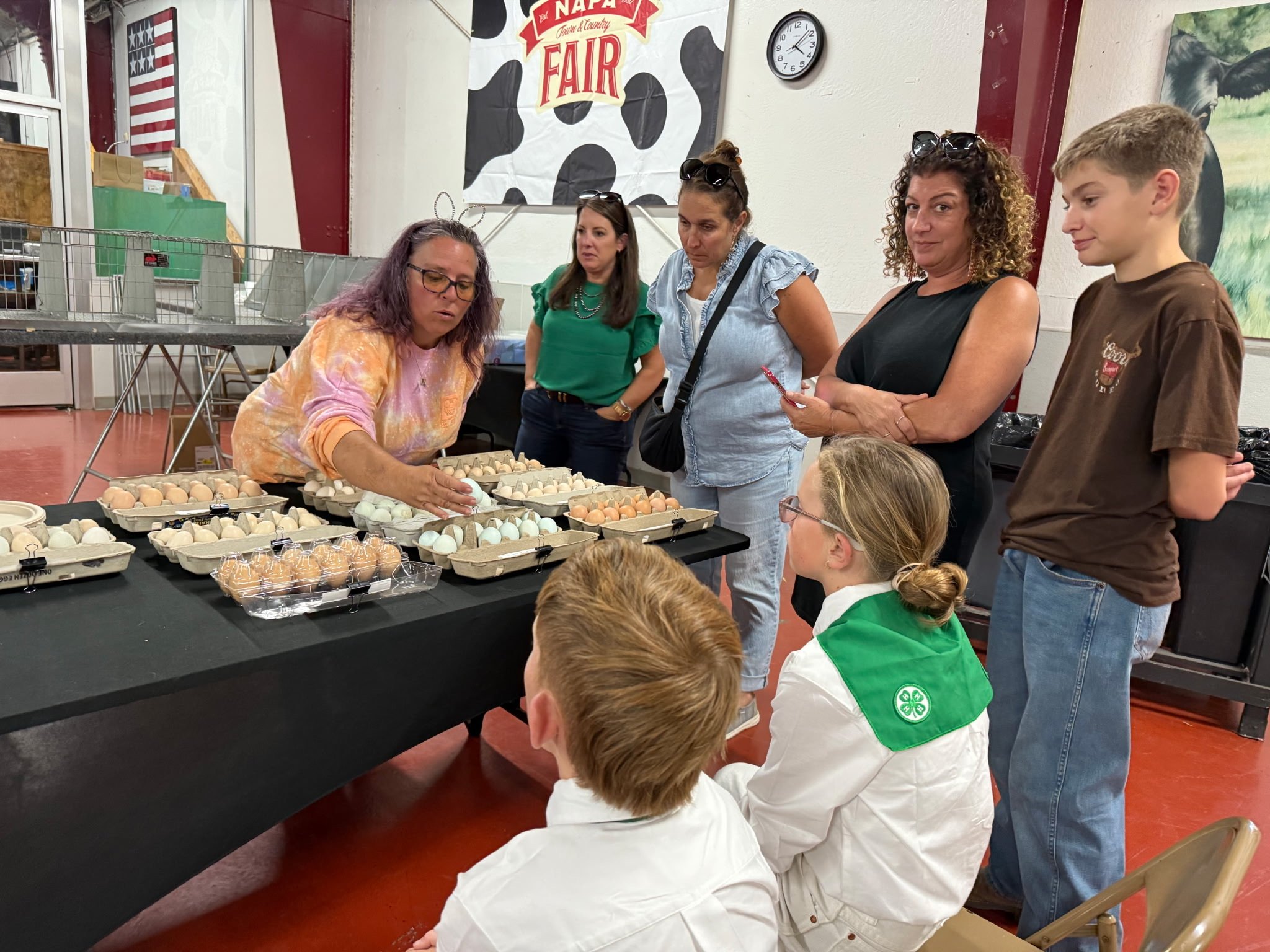 A woman demonstrating eggs to a group of children and adults at a fair. The eggs are arranged on a table in trays, and the setting appears to be an indoor community event space with a farm-themed banner in the background.