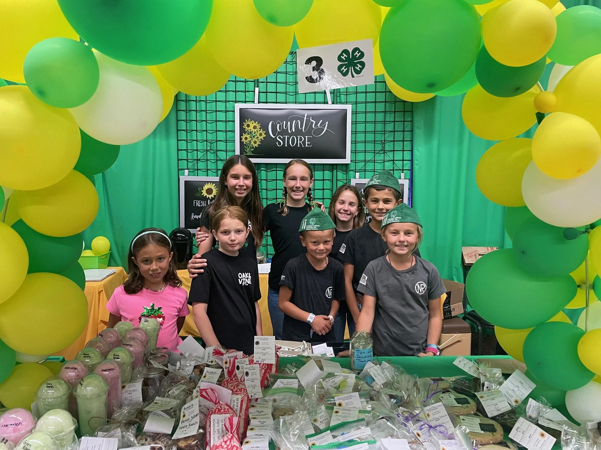 Group of children dressed in scout uniforms and casual clothes standing behind a table at a country store booth decorated with green and yellow balloons.