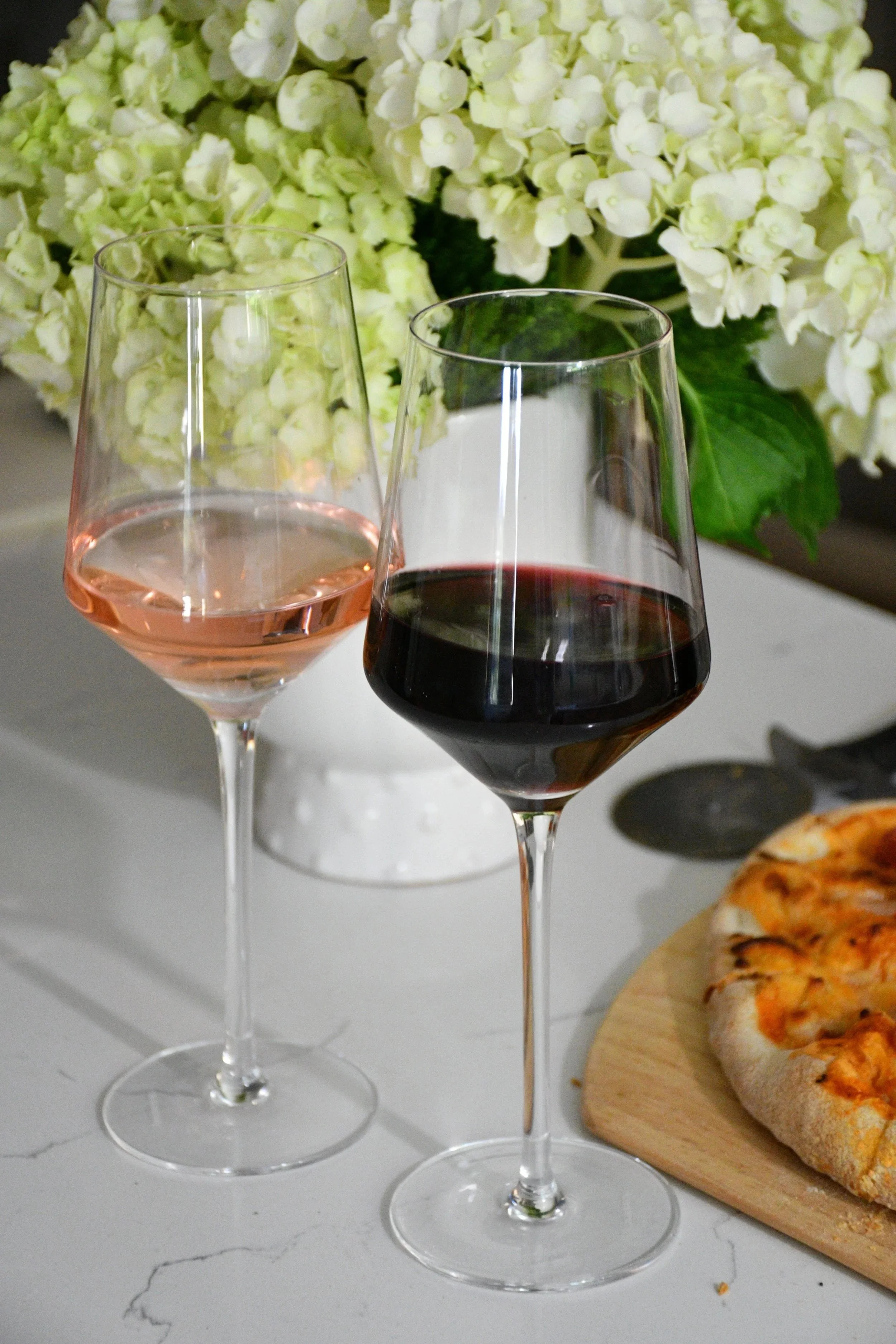 Two glasses of wine, one with rosé and the other with red wine, on a white marble table with a pizza and a bouquet of white flowers in the background.