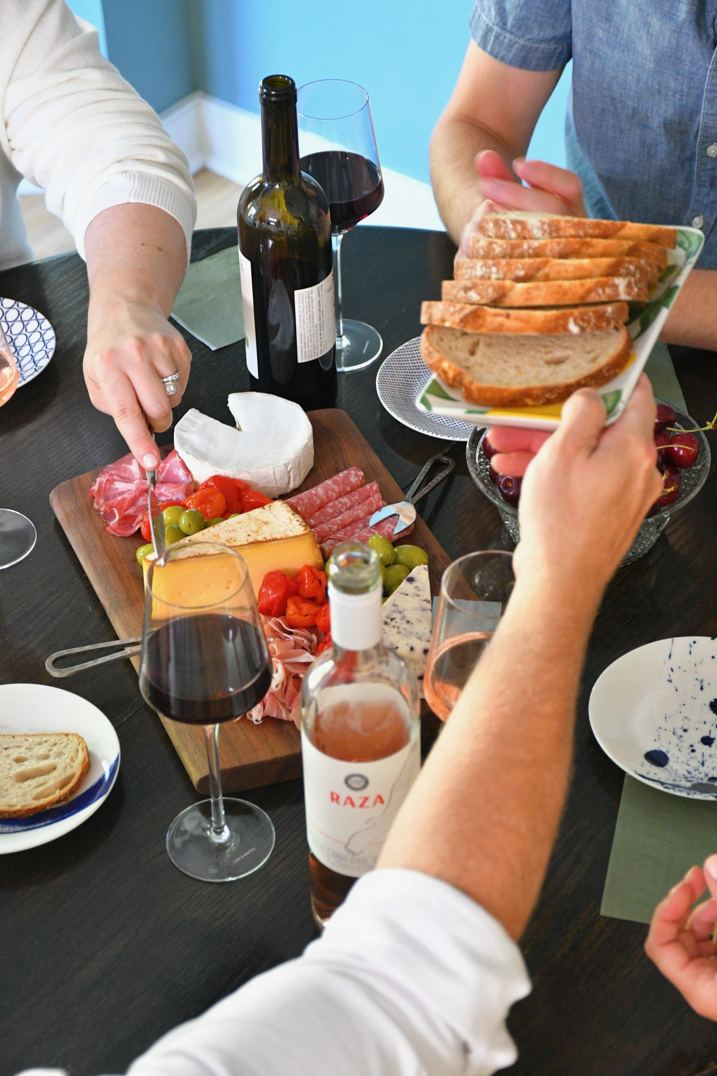 People serving sliced bread and various cheeses, meats, and grapes on a table set for a meal with wine glasses and bottles.