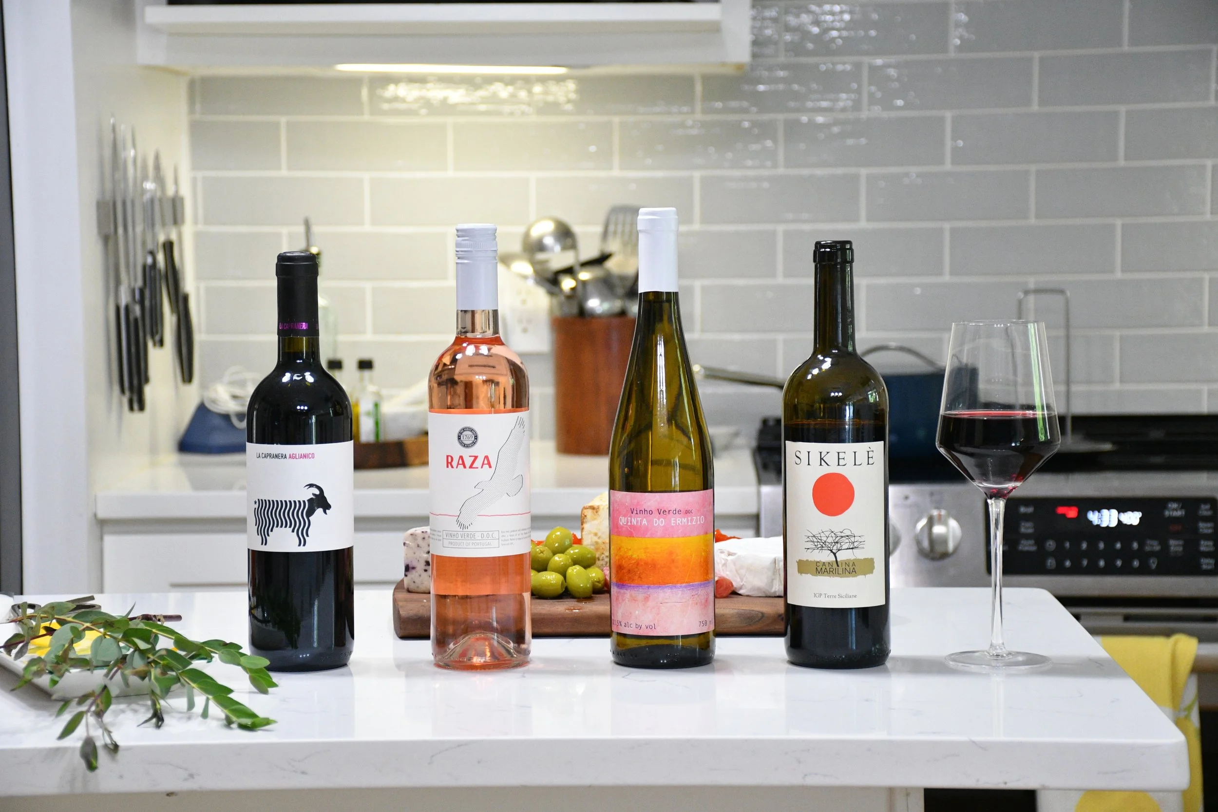 Five bottles of wine lined up on a white kitchen counter, with a wine glass filled with red wine on the right. In the background, there is a cutting board with green olives and cheese, a knife block, and kitchen utensils.