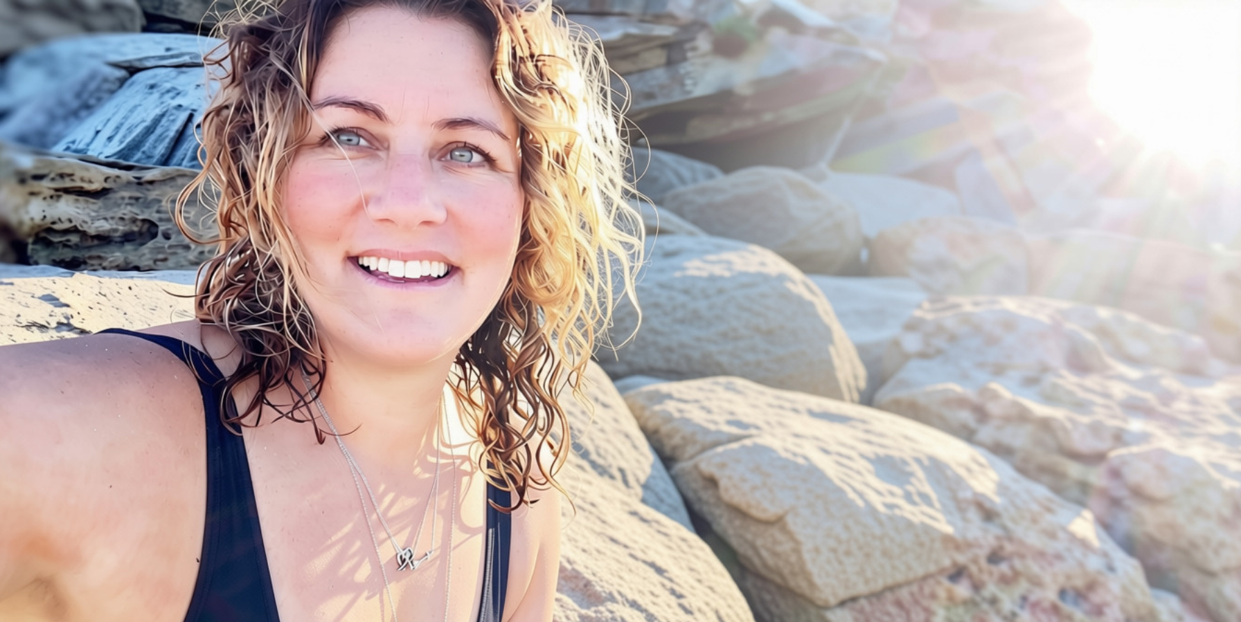 Close-up of a smiling woman with curly blonde hair, wearing a black tank top and silver jewelry, sitting on rocks with sunlight and rock formations in the background.