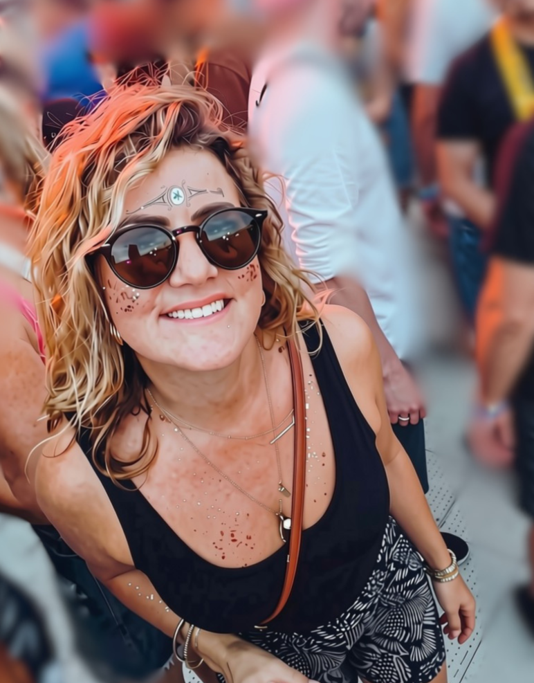 Smiling woman with sunglasses and face paint, at a crowded outdoor event.
