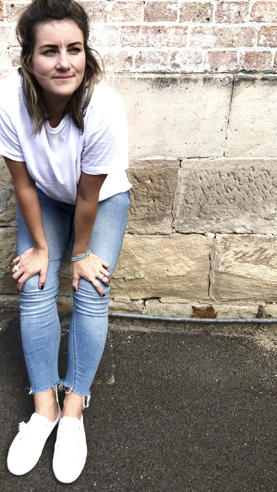Person wearing a white t-shirt, blue jeans, and white sneakers, standing against a stone wall.