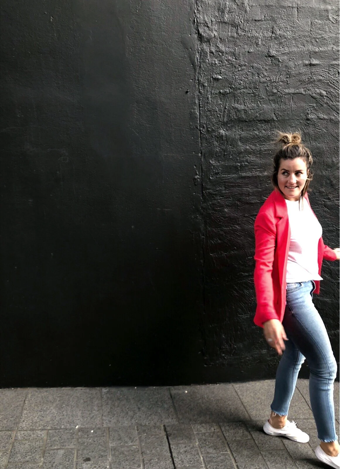 Woman in red jacket and jeans walking beside a black wall.