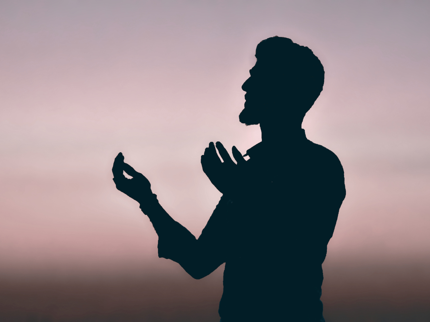 Silhouette of a man praying or meditating against a soft pink and gray sky.