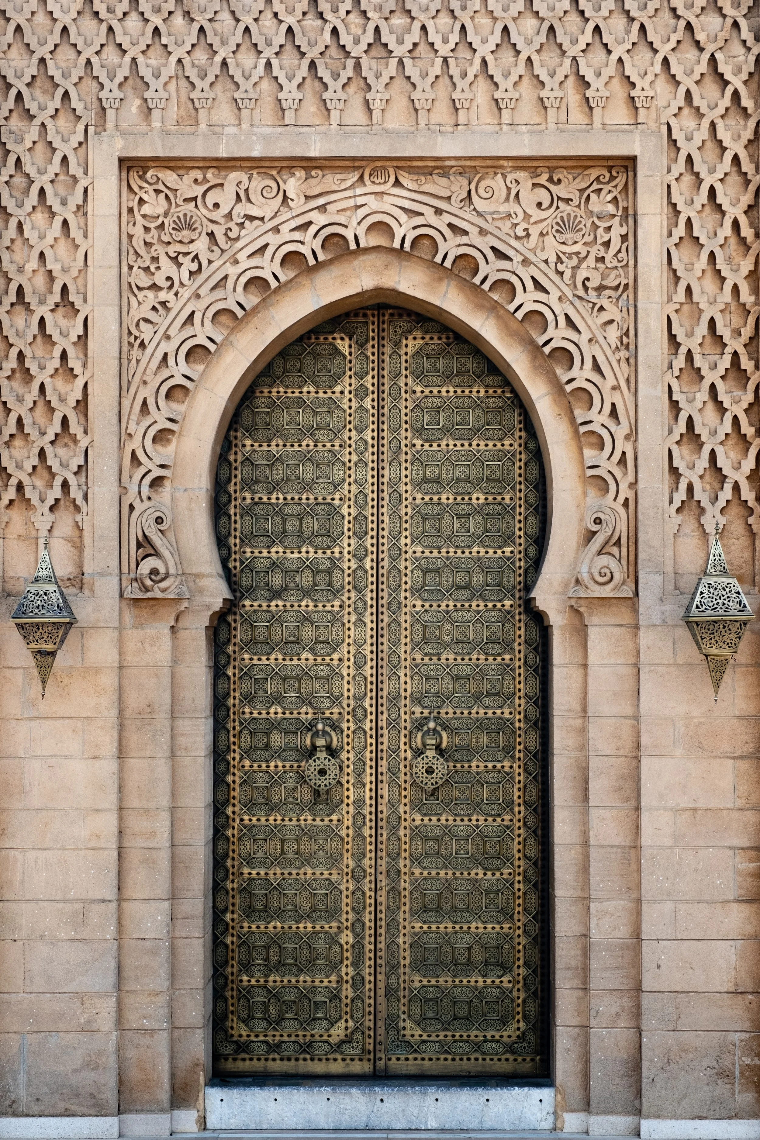 Ornate metal double doors set within an arched stone doorway, surrounded by intricate Arabic-style geometric and floral carvings, with two lantern-style wall sconces on either side.