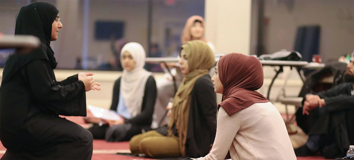 A woman in a black hijab and black outfit is speaking to a group of women sitting on the floor, all wearing hijabs and listening attentively.