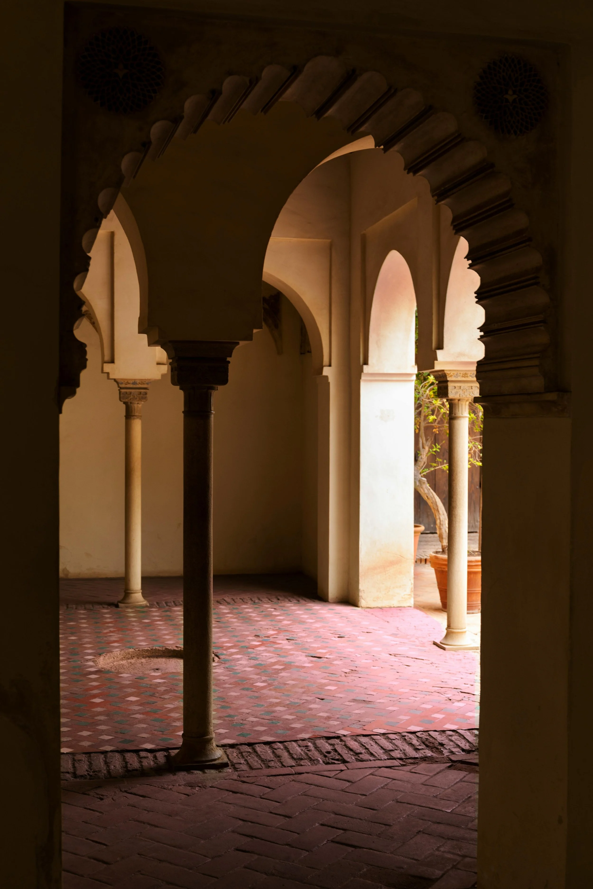 Archways and columns in a courtyard with pink and red brick flooring and potted plants.