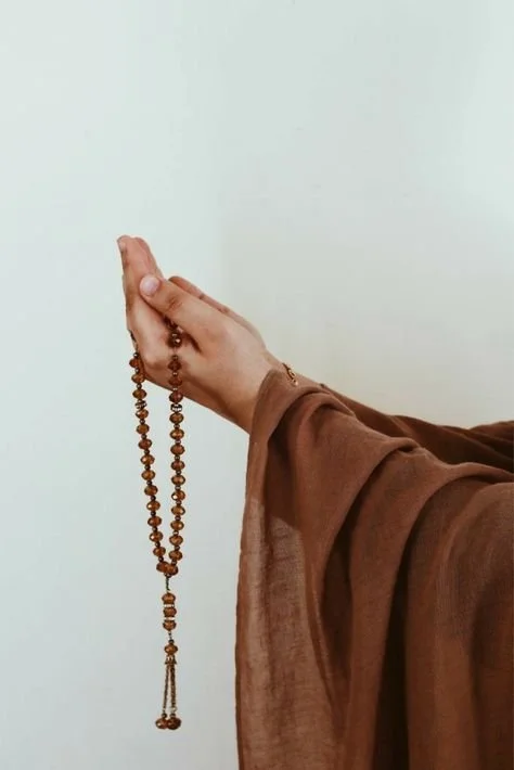 A person wearing brown clothing holding a wooden prayer bead necklace in front of a plain light-colored background.