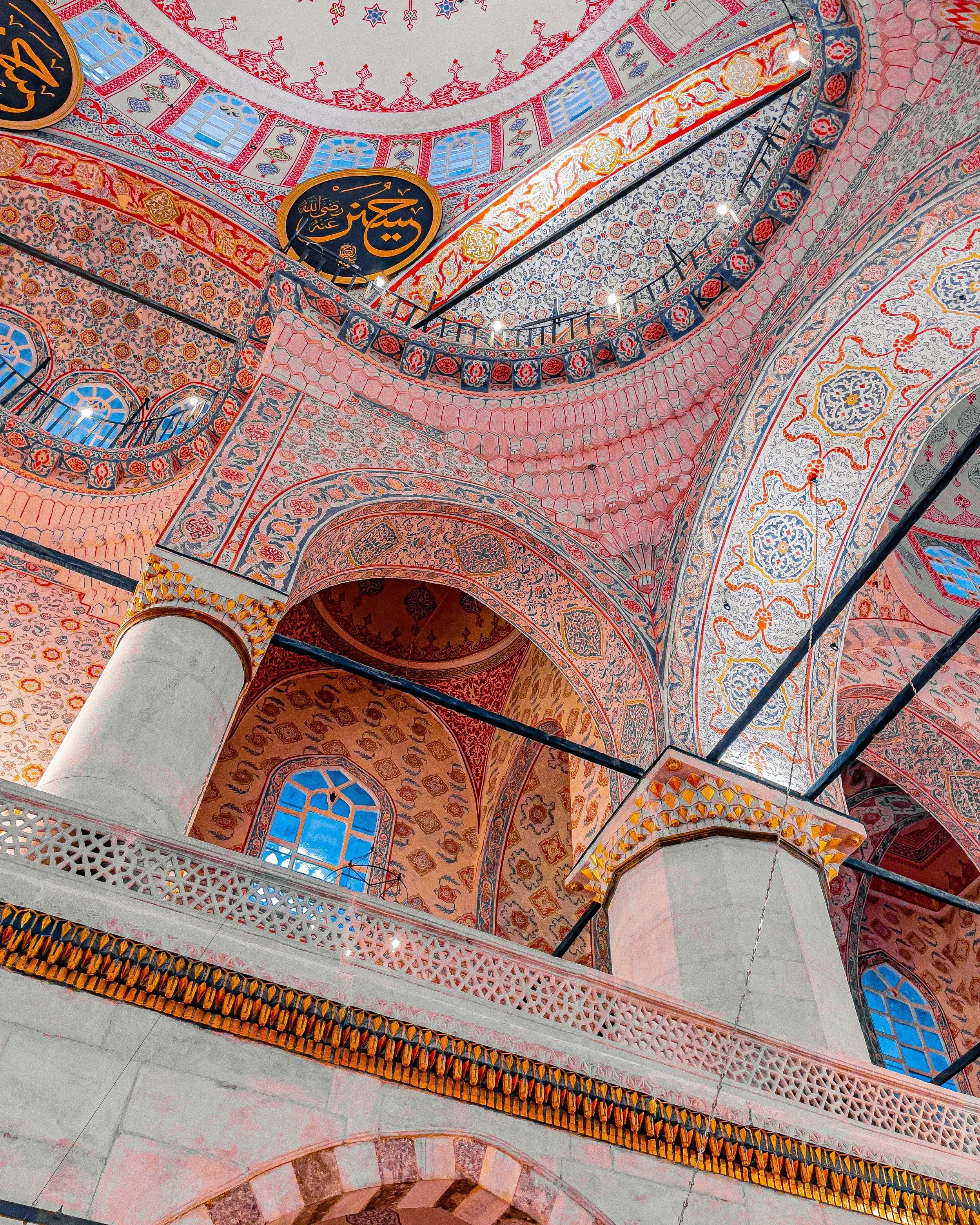 Interior view of a mosque with ornate, colorful Islamic tile designs on the walls and arches, large columns supporting the structure, and Arabic calligraphy on circular plaques.